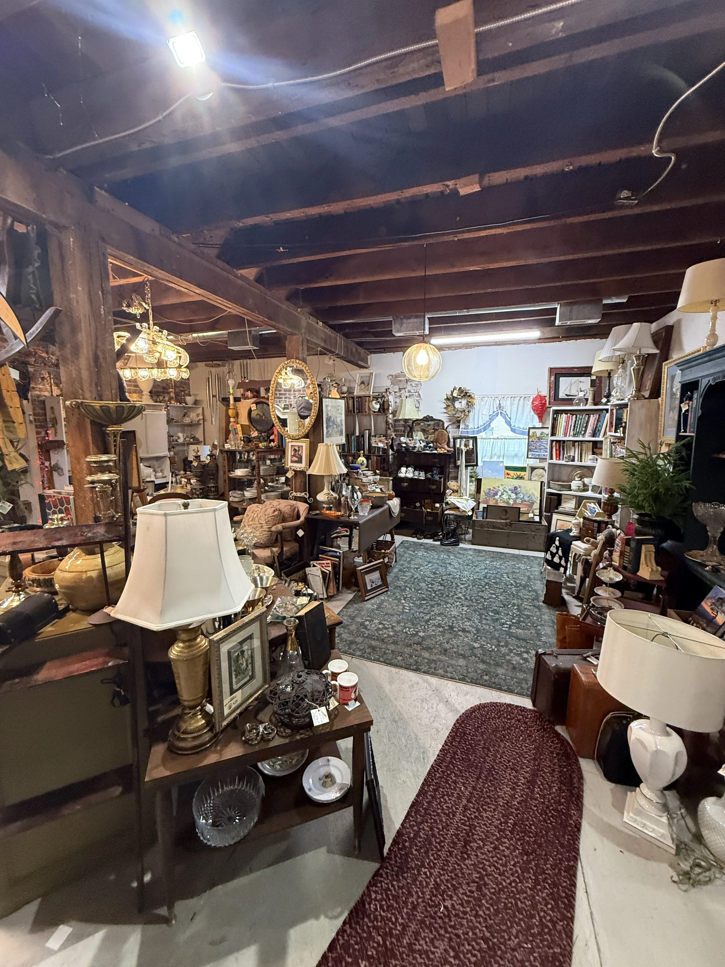 Cluttered antique shop interior filled with vintage furniture, lamps, books, paintings, mirrors, dishes, and decorative objects beneath exposed wooden ceiling beams.