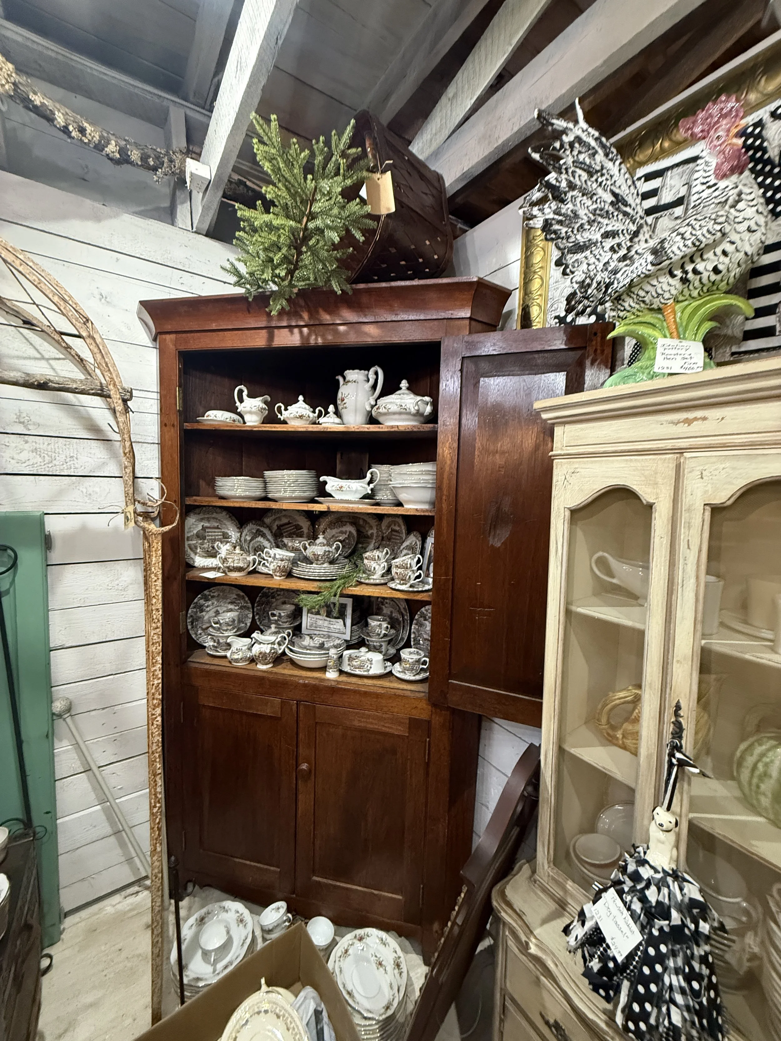 Antique wooden china cabinet displaying vintage teacups, saucers, plates, and serving pieces in a collectibles shop