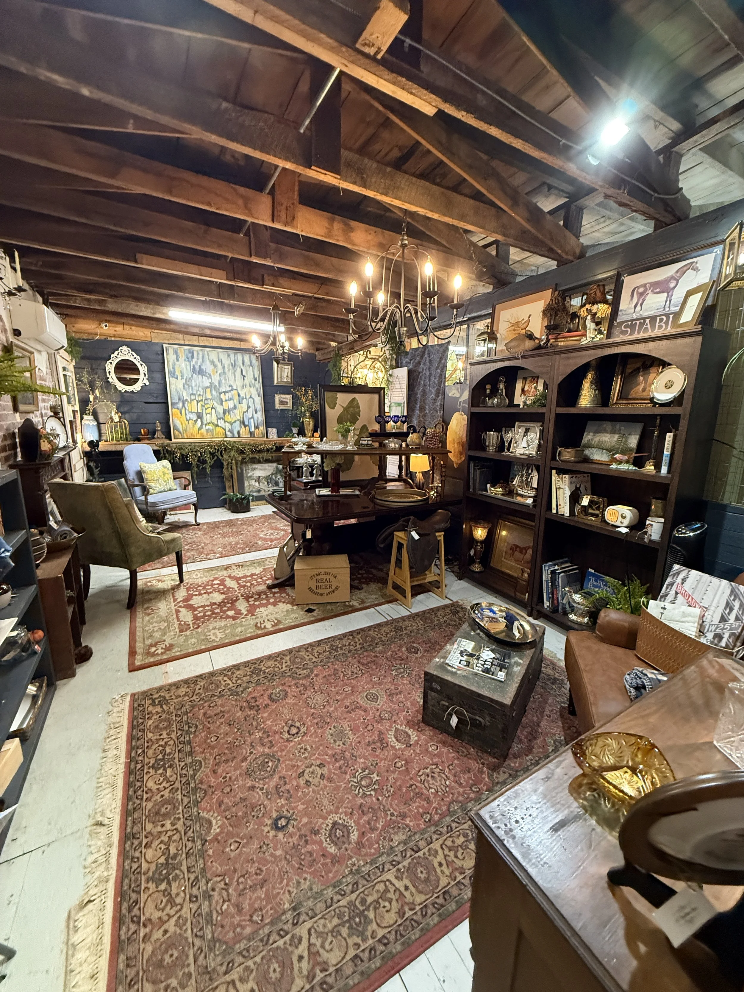 Eclectic antique shop interior with vintage furniture, artwork, books, rugs, collectibles, chandeliers, and exposed wooden ceiling beams