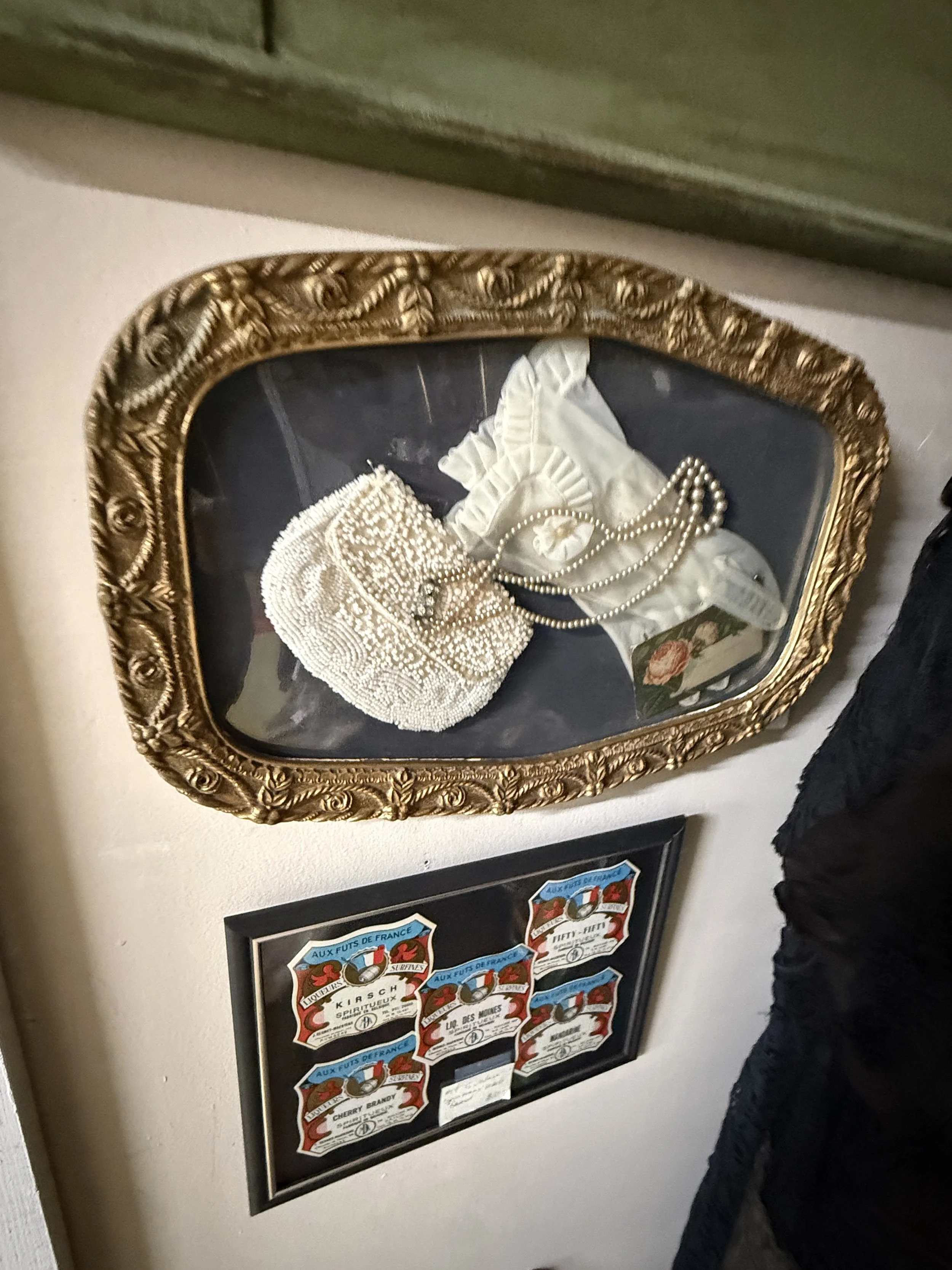 Ornate gold-framed display tray holding a beaded clutch, white fabric, jewelry, and a floral accessory above framed vintage French liquor labels