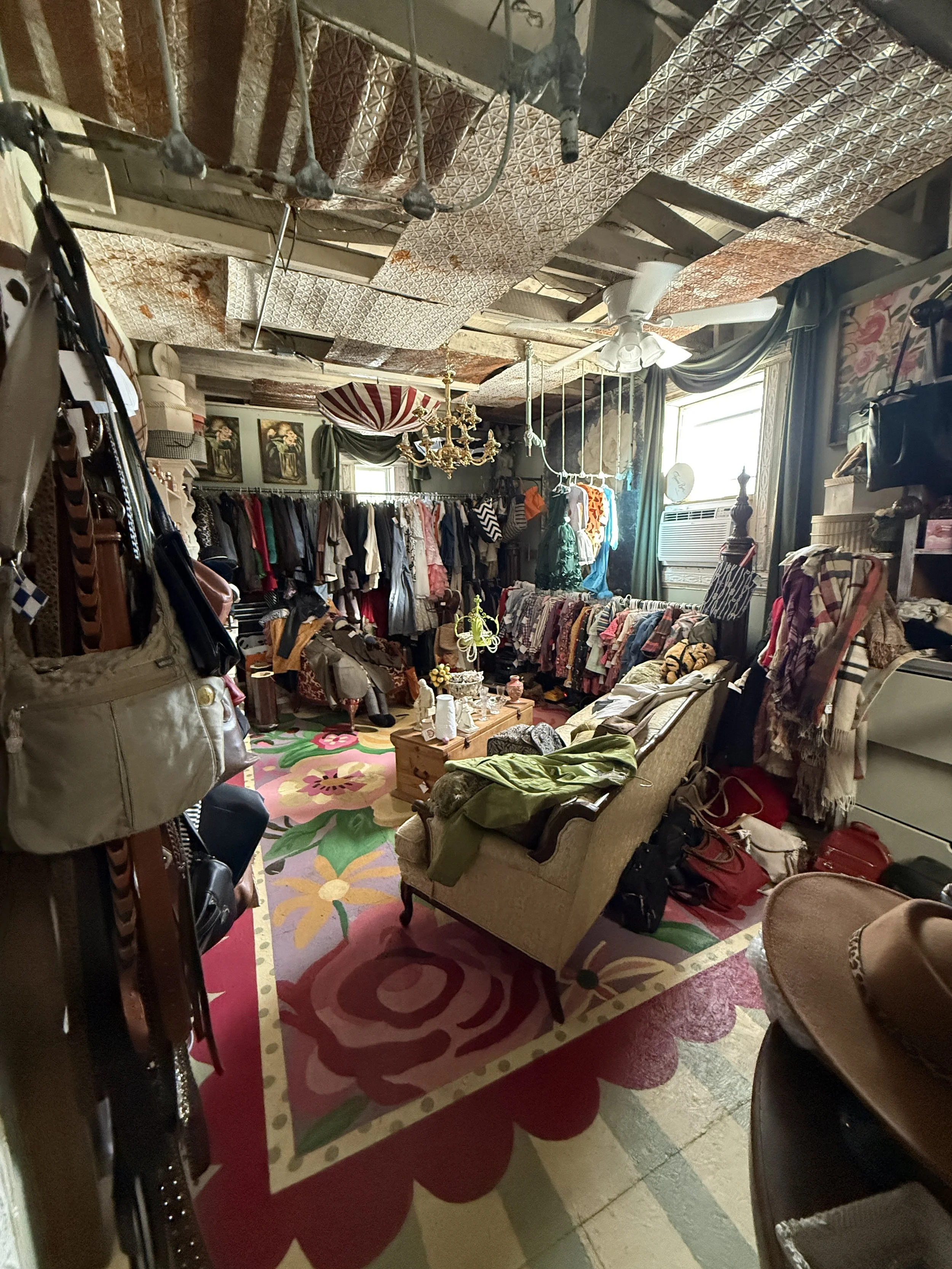 Eclectic vintage clothing shop interior with racks of garments, colorful floral rug, ornate chandelier, ceiling fan, and reflective insulated ceiling panels