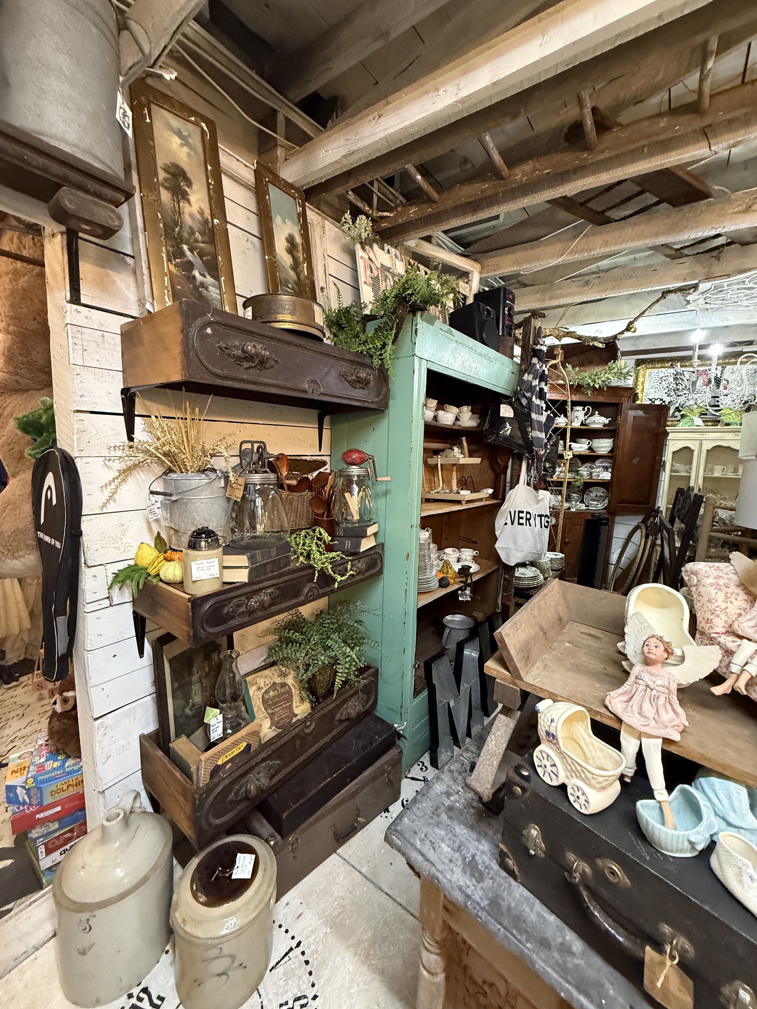 Vintage antique store display filled with antique furniture, framed artwork, dishes, plants, toys, and assorted home decor beneath exposed wooden beams.