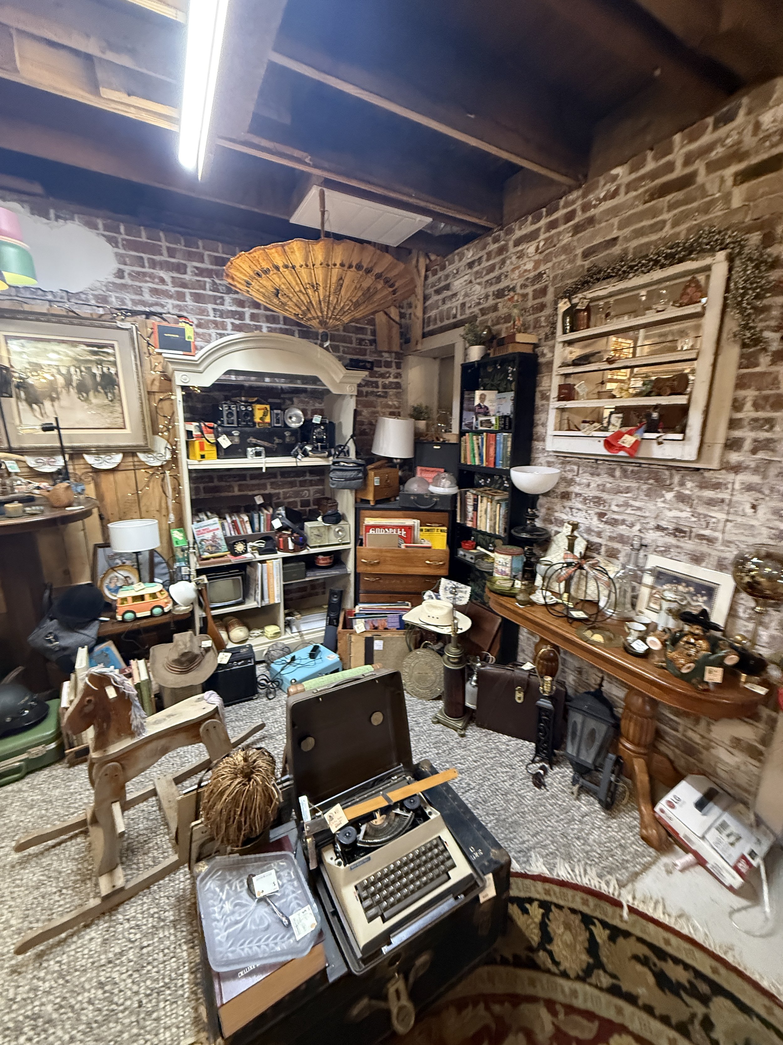 Vintage antique shop interior with exposed brick walls, shelves of collectibles, an old typewriter, books, furniture, lamps, and decorative objects.