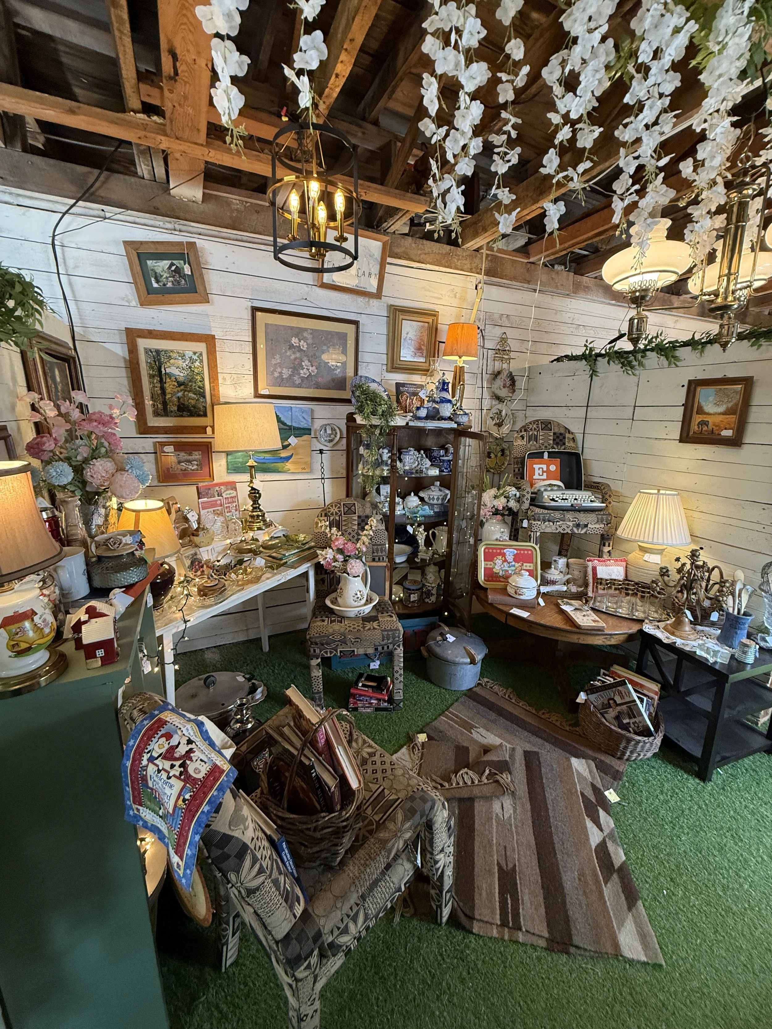 Eclectic antique shop interior with framed artwork, vintage lamps, dishes, furniture, baskets, and hanging white flowers beneath exposed wooden beams
