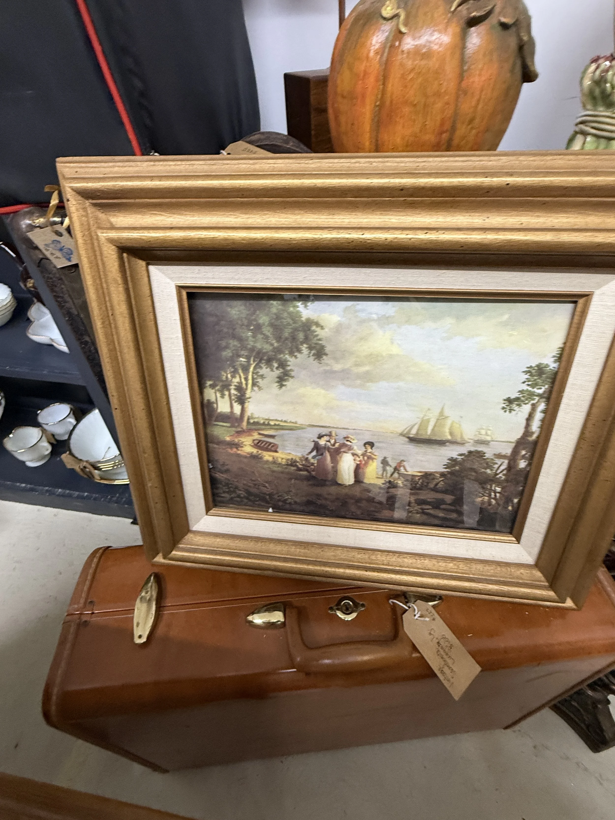 Ornate wooden-framed vintage landscape painting depicting people and sailboats by a waterfront, displayed on an antique wooden table