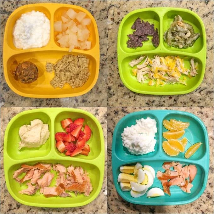 Four divided plates with different food items: yellow plate with rice, diced melon, a meatball, and crackers; green plate with grape leaves and shredded chicken salad; another green plate with mashed potato, diced strawberries, and shredded cooked sa