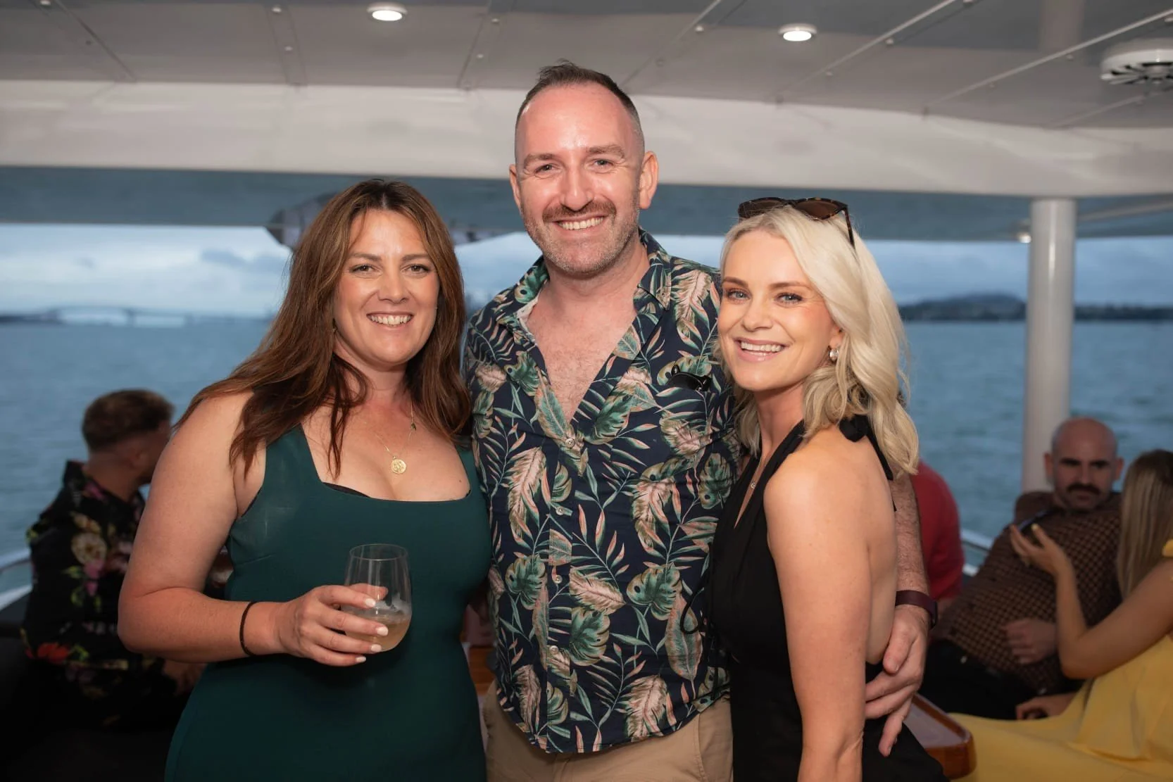 Three people smiling and posing together on a boat, with water and a bridge in the background, during a social event.
