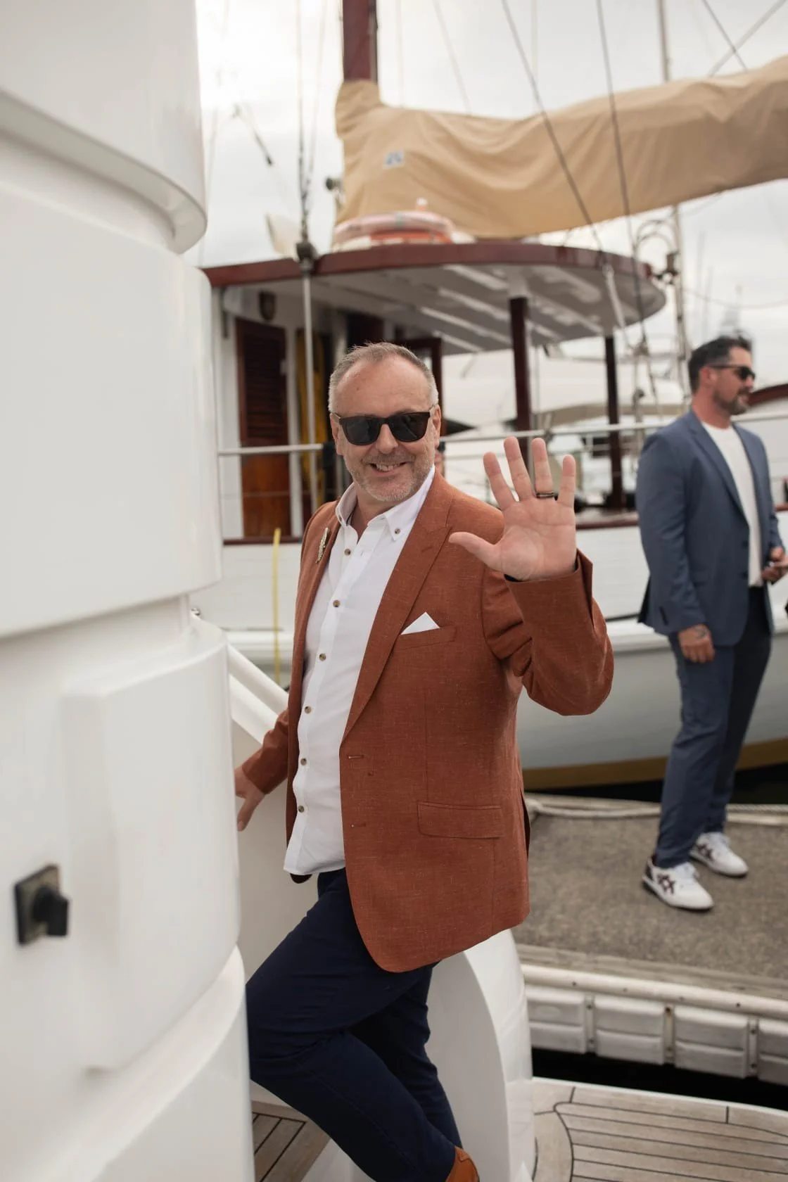 A man with sunglasses, a brown blazer, and a white shirt smiling and waving on a yacht.
