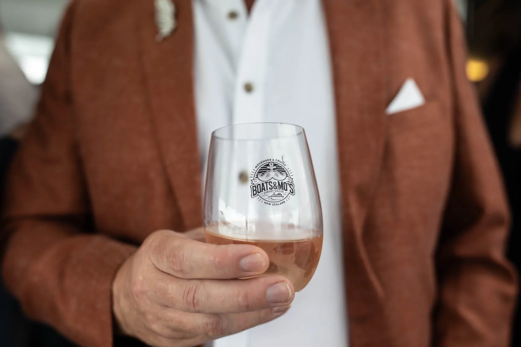 Person holding a wine glass with pink beverage, the glass has 'Boats & M0's' logo, person wearing a white shirt and a brown jacket, blurred background.