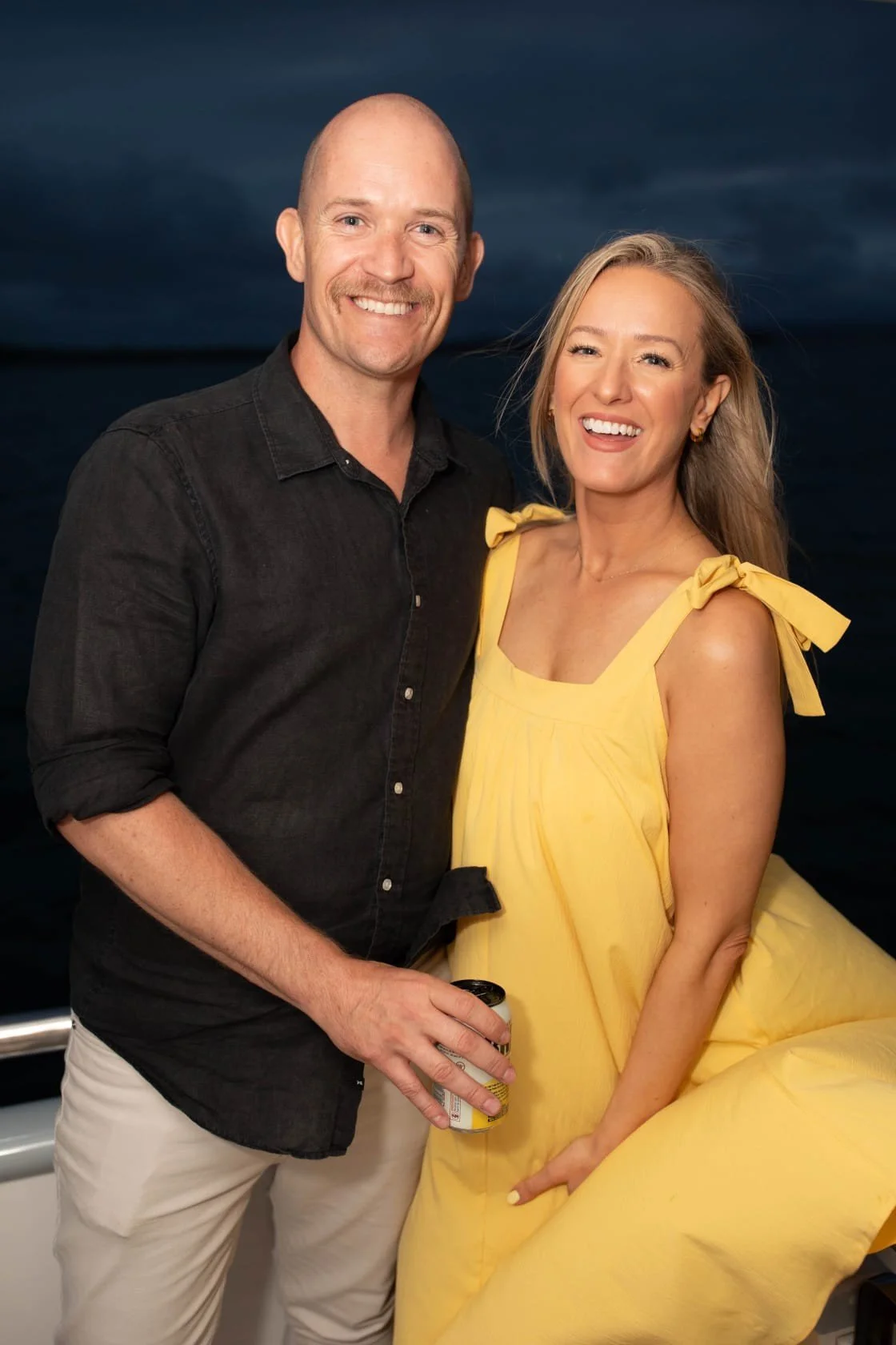 A man and woman smiling on a boat, with a dark ocean background, the man in a black shirt and the woman in a yellow dress