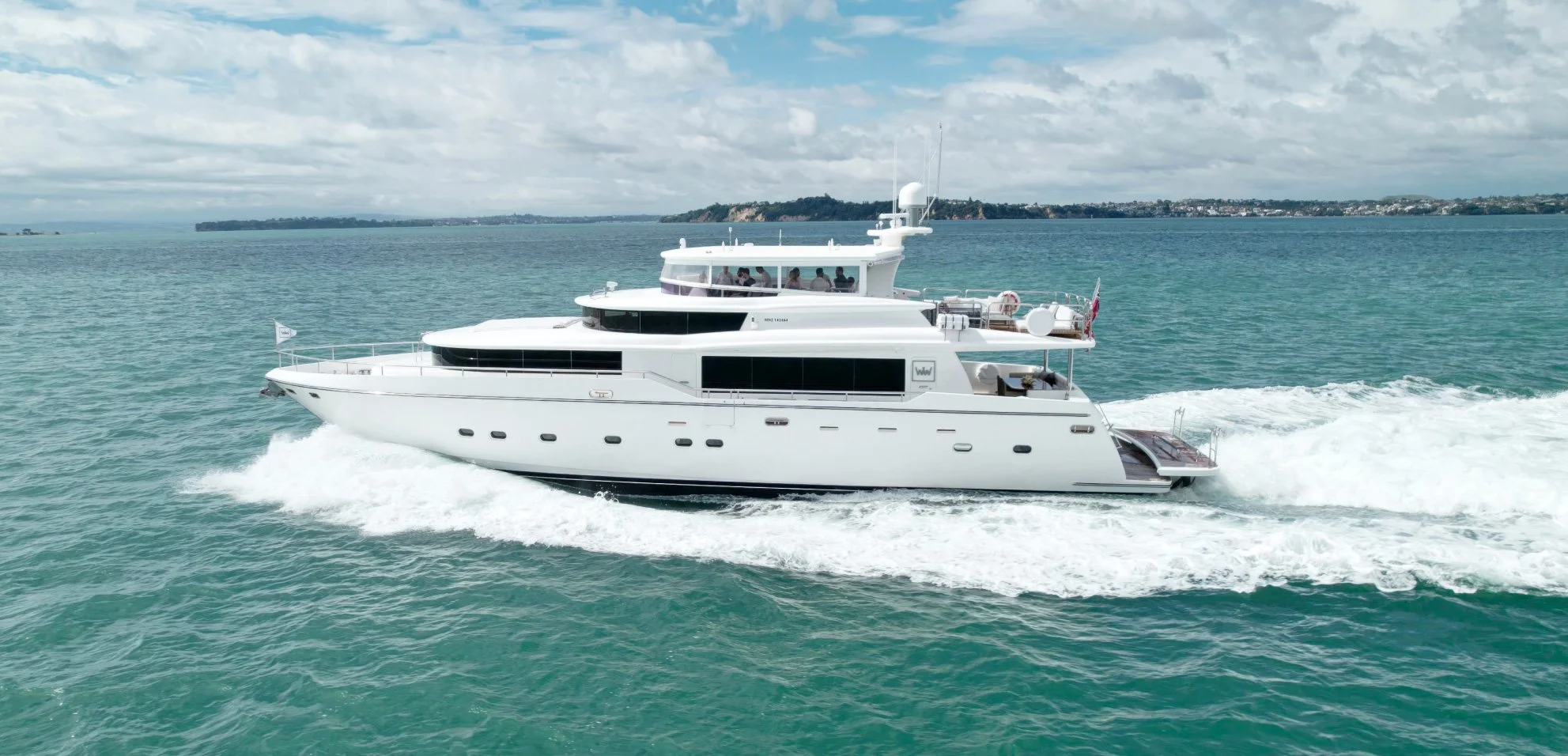 A large white yacht cruising through the water with a group of people visible on the upper deck, water and a distant shoreline under a partly cloudy sky.