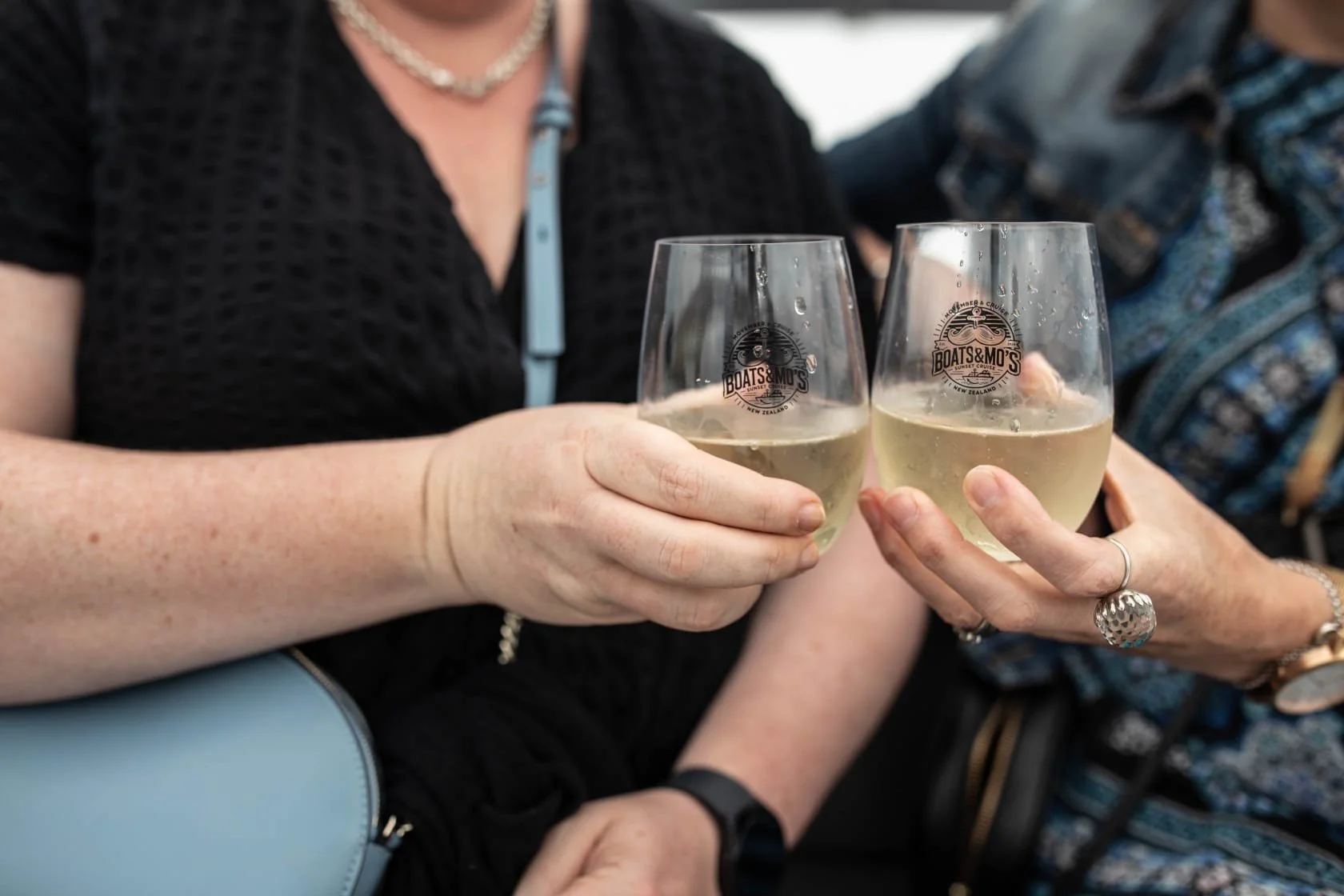 Two women clinking glasses of white wine, with one wearing rings and jewelry, and a woman in a black dress, at a social gathering.