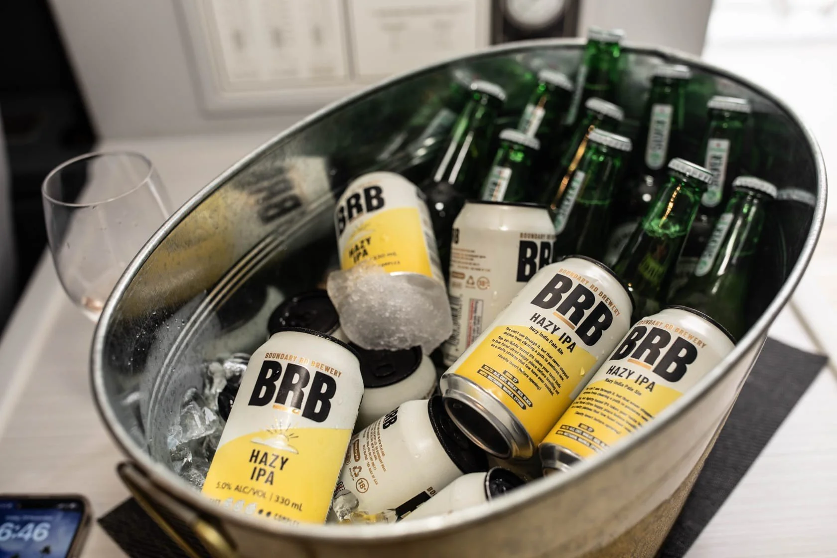 A metal tub filled with ice and several cans of BRB Hazy IPA beer, some green glass bottles, an empty glass, and bottle caps.