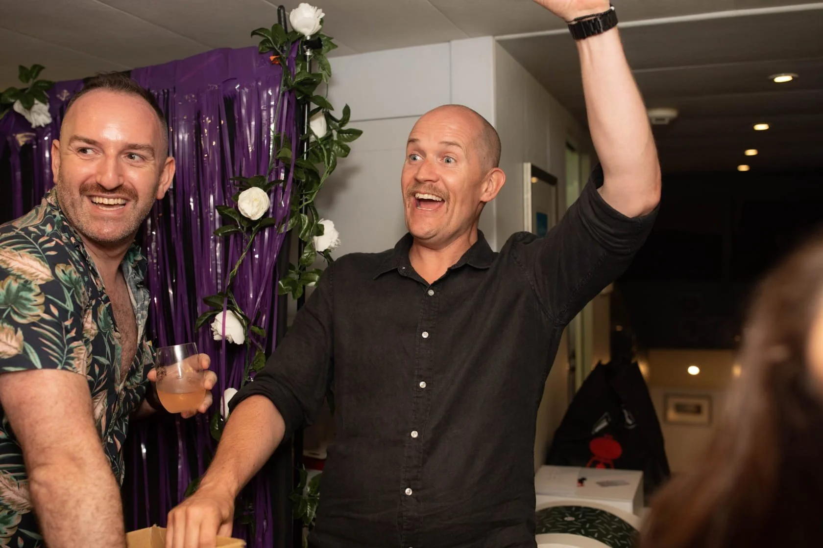 Two men smiling and celebrating at a party, one holding a glass of drink, with purple decorations and white flowers in the background.