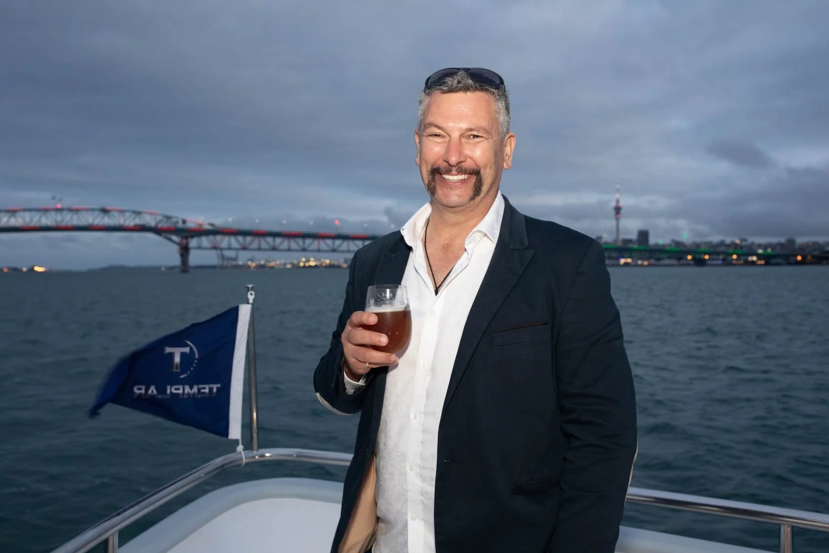 A man in a dark suit jacket and white shirt standing on a boat at dusk, holding a glass of beer, with a city skyline and a bridge in the background.