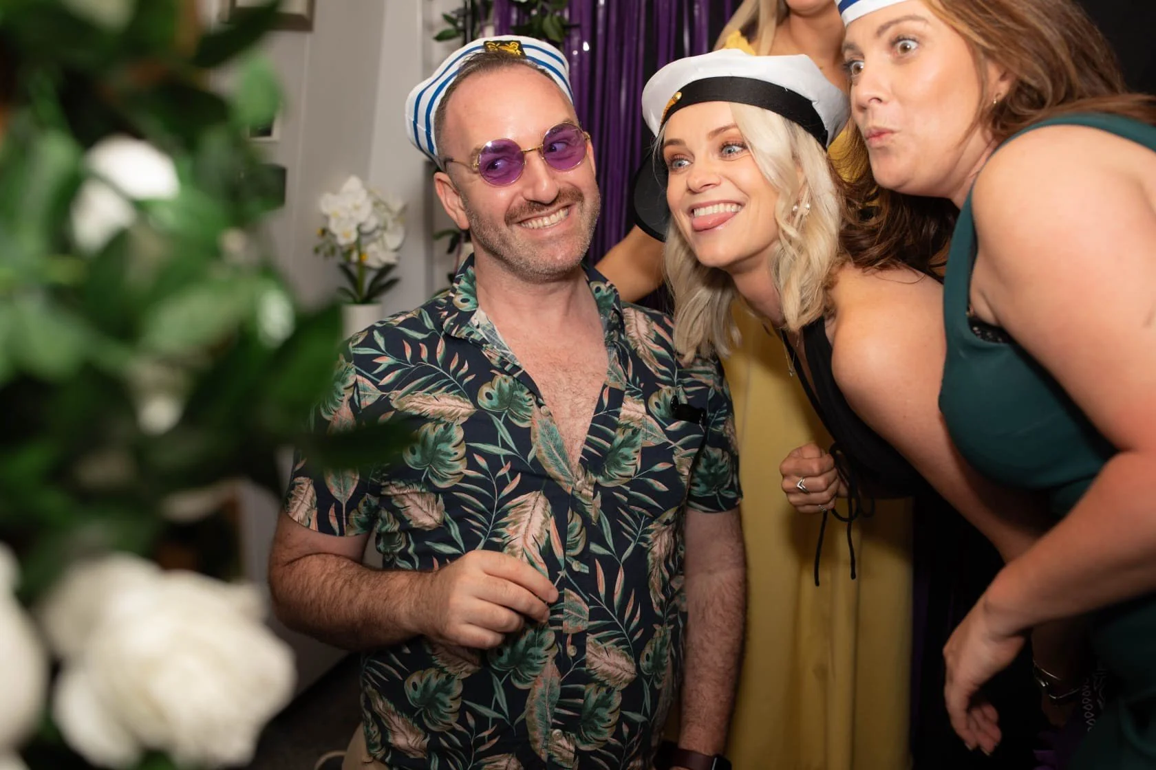 Three people at a party smiling and posing for a photo, dressed in fun beach-themed costumes, with a backdrop of purple and yellow curtains and white flowers.