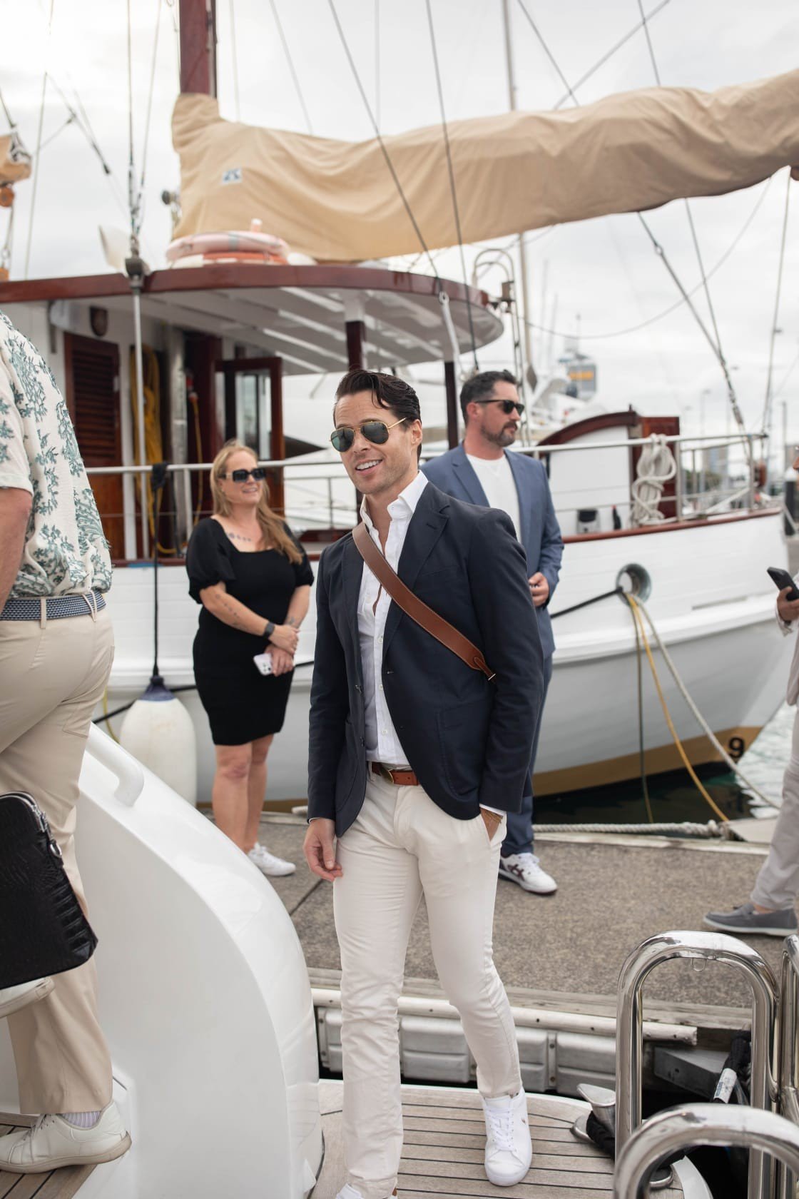 Group of five people on a yacht dock, with a man in a navy blazer and white pants in the foreground, smiling and wearing sunglasses.