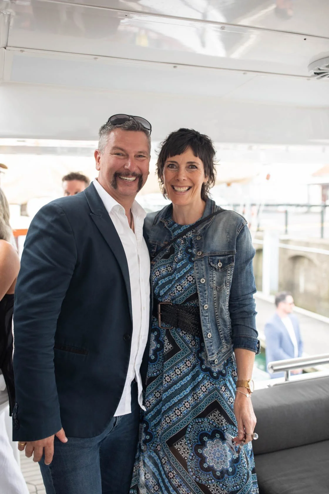 A man and woman smiling and posing together on a boat or yacht with water and a man in a suit in the background.