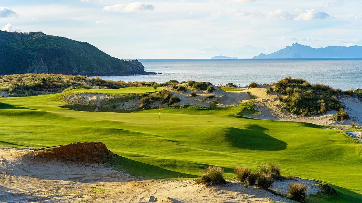 Golf course with green grass, sand dunes, and shrubs, located along a coastline with mountains in the background. Te Arai Links, ranked top 40 courses in the world.