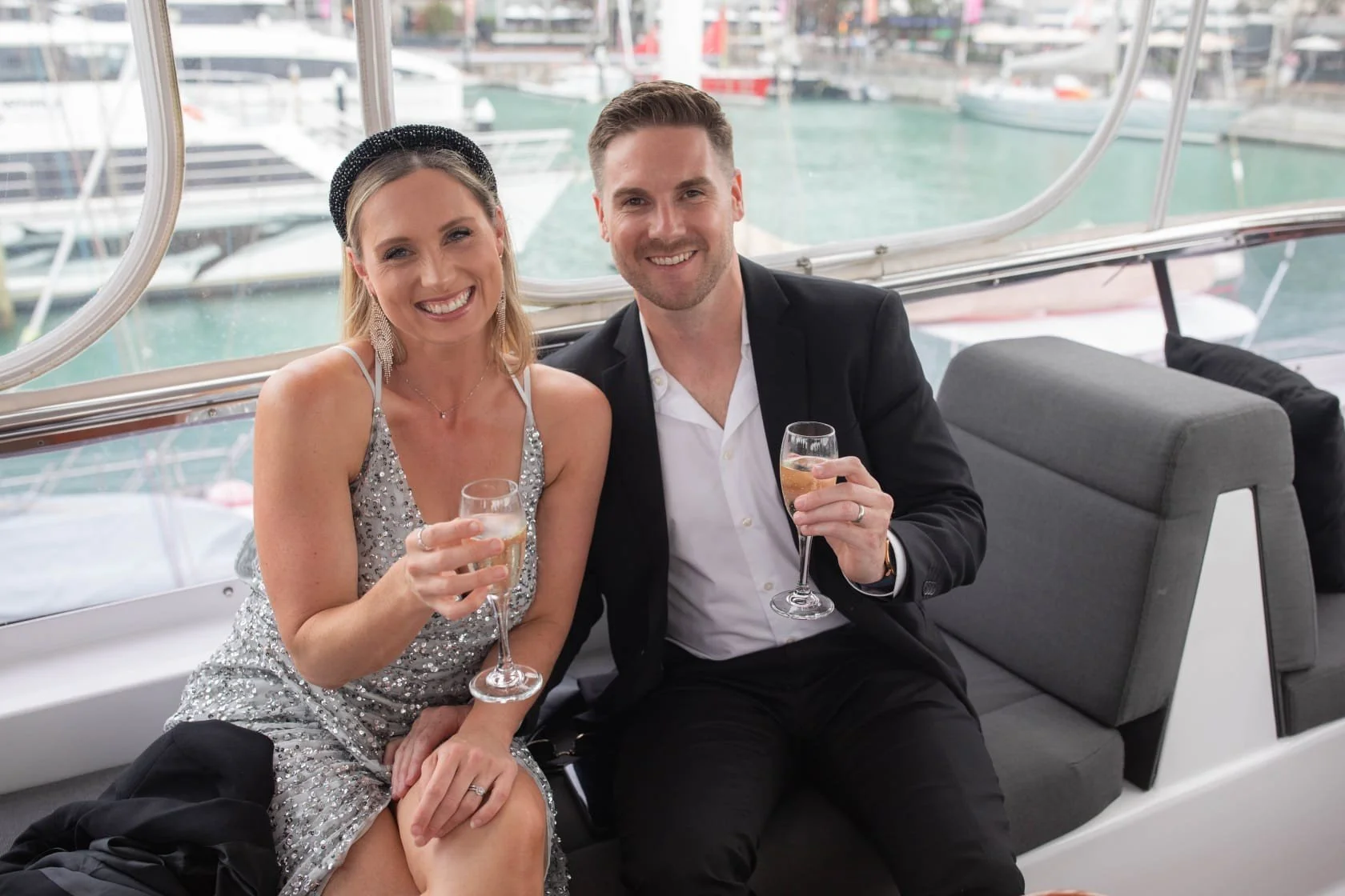 A smiling man and woman dressed in formal attire sitting on a boat with a marina and yachts in the background, holding glasses of champagne.
