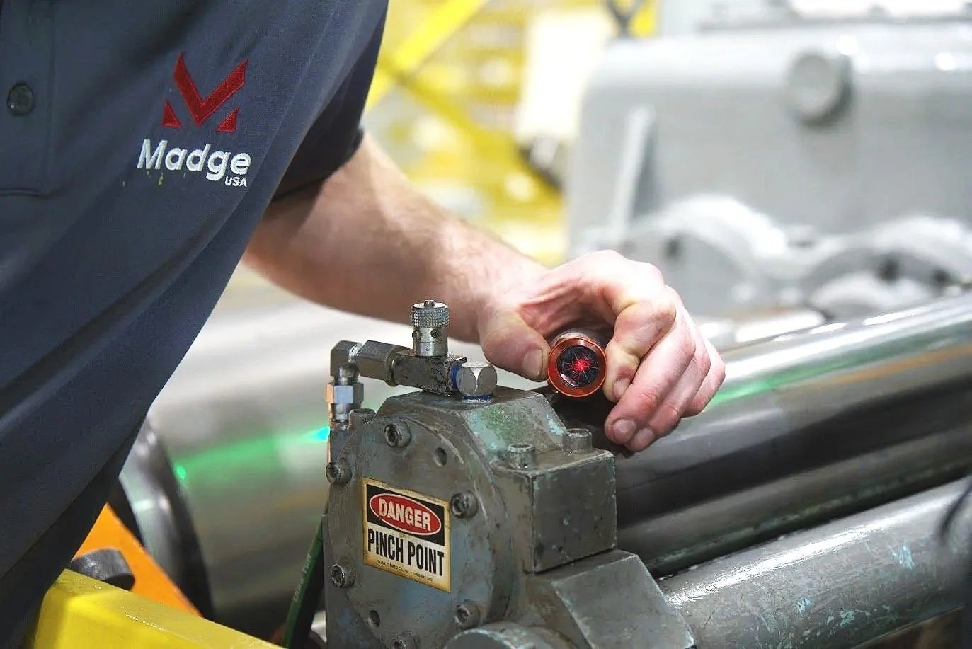 A worker in a dark uniform with a Madge logo inspects a device with a flashlight in an industrial setting, with a yellow safety rail and machinery in the background.