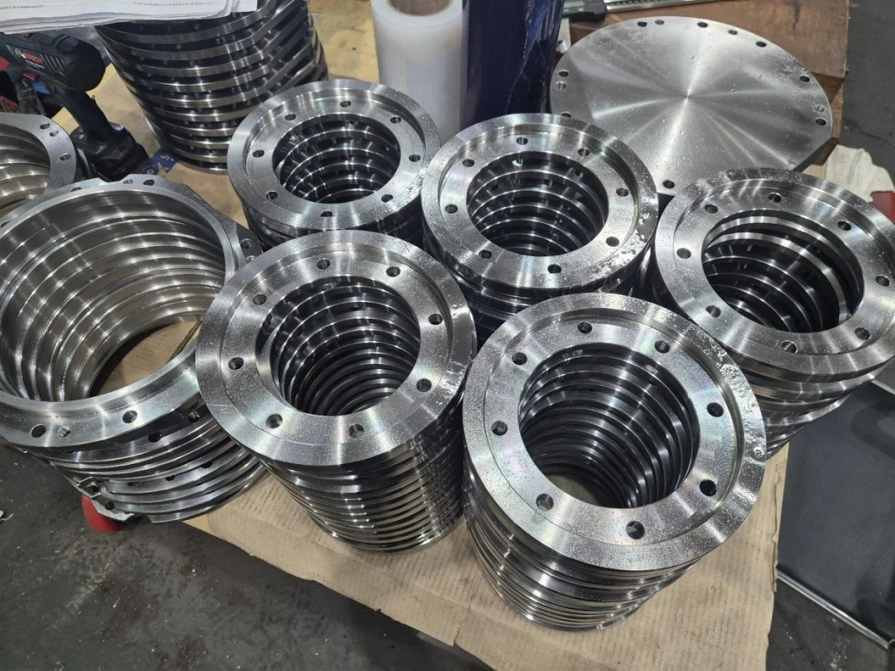 Multiple metallic industrial flanges stacked on a workbench in a factory setting.