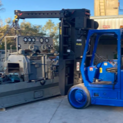 Blue forklift with black forks beside a gray truck with equipment and a ladder, in an outdoor industrial area with trees and other vehicles in the background.
