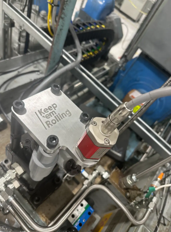 Close-up of industrial machinery with a metal plate that says 'Keep Em Rolling' attached to a cylindrical component, surrounded by various metallic pipes and electrical wiring in a factory setting.