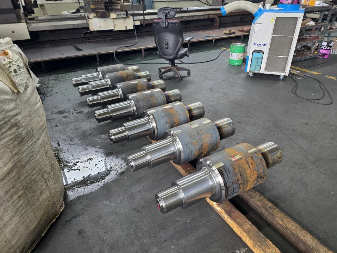 Row of large metal industrial rollers or shafts on the floor of a workshop or factory, with various tools and equipment in the background.
