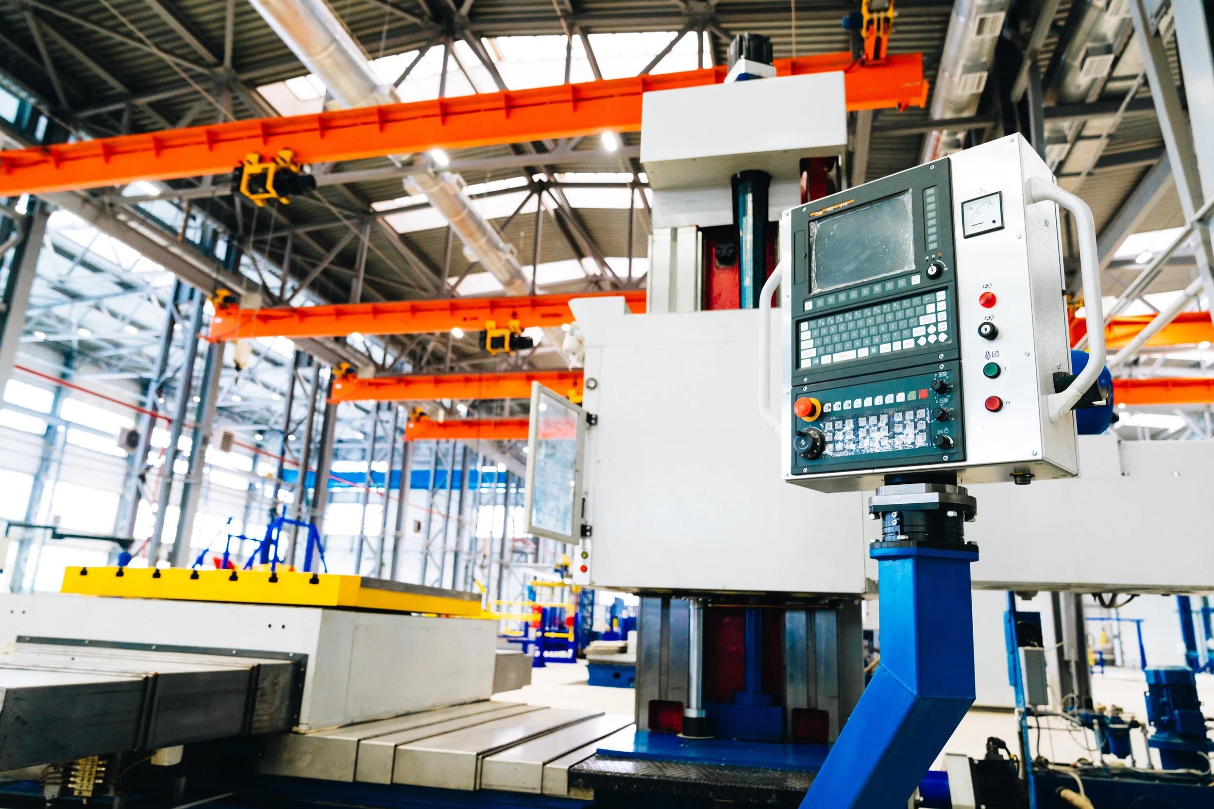 Industrial manufacturing equipment with a control panel and orange overhead cranes in a factory setting.