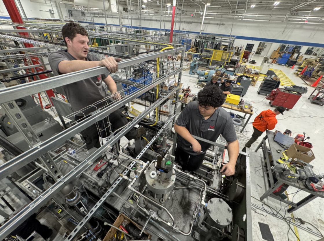 Workers in a manufacturing or industrial setting assembling or maintaining machinery, with various tools and equipment around them.