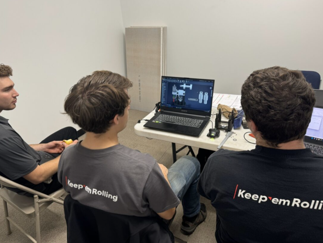 Three young men sit at a table looking at a laptop with a CAD software project displayed, surrounded by tools and papers, in a room with a plain wall.