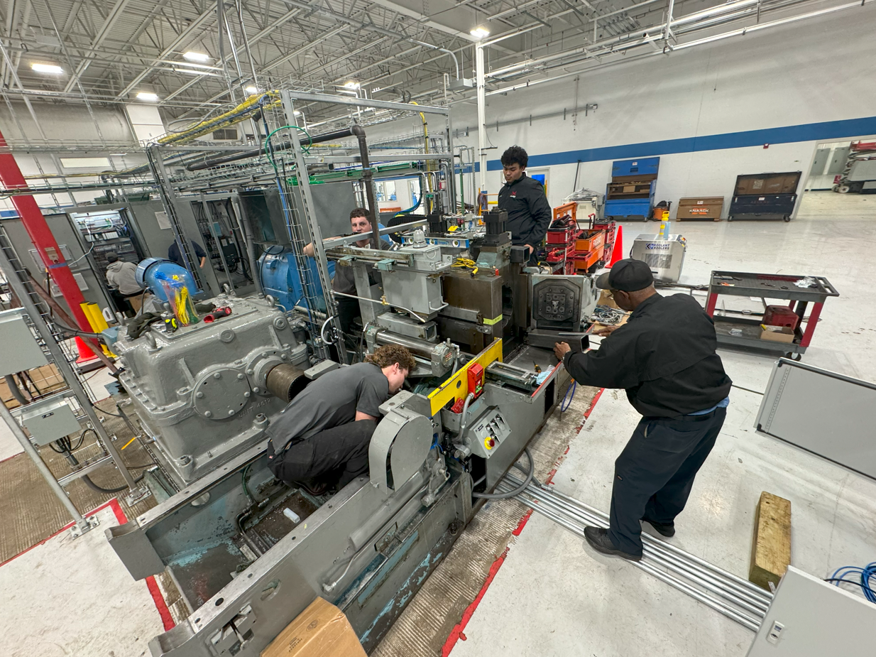 Four workers inspecting and working on large industrial machinery inside a manufacturing facility.