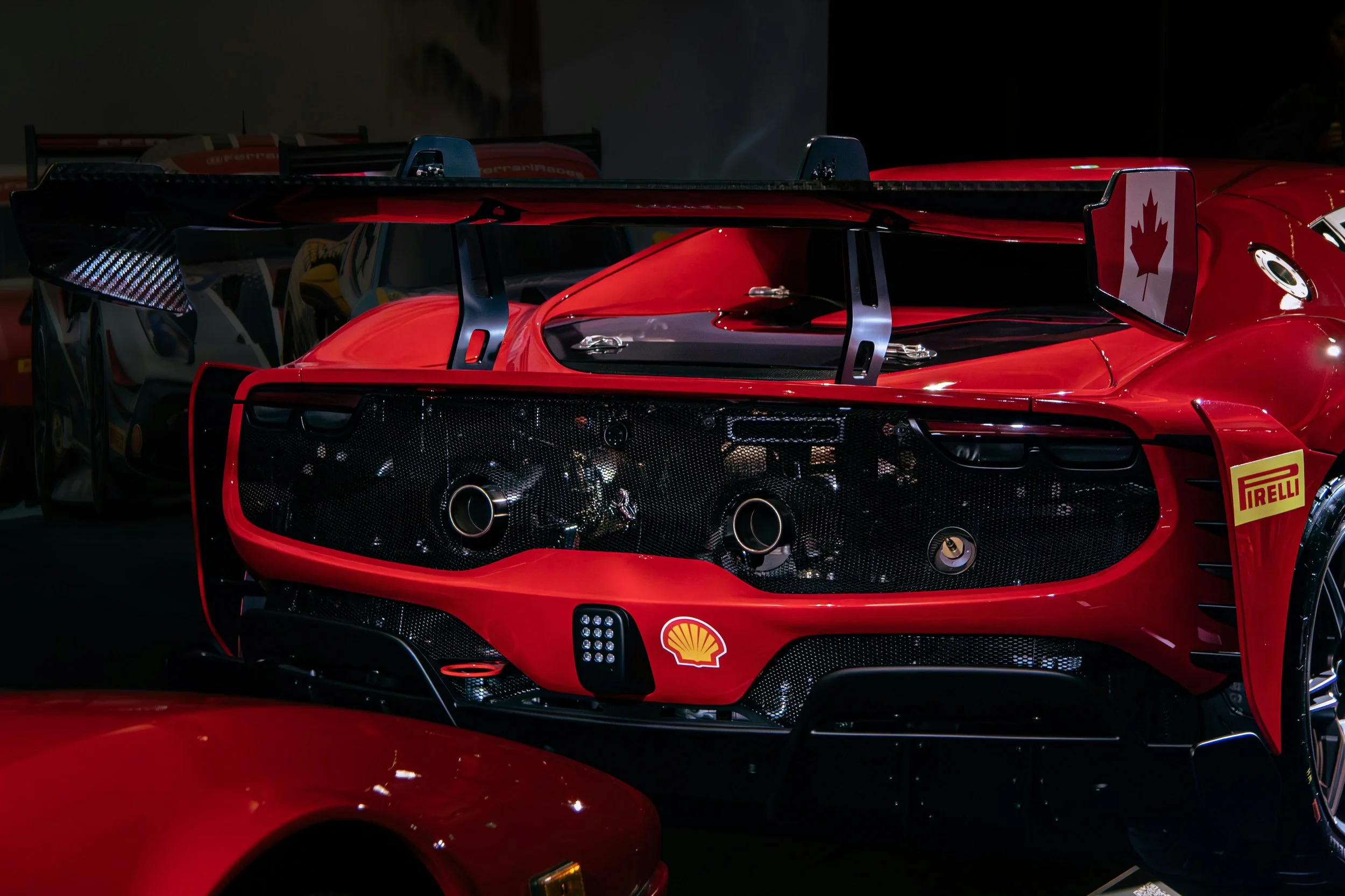 Red racing car with a large rear wing and sponsor decals, including Pirelli, Shell, and a Canadian flag, displayed indoors.