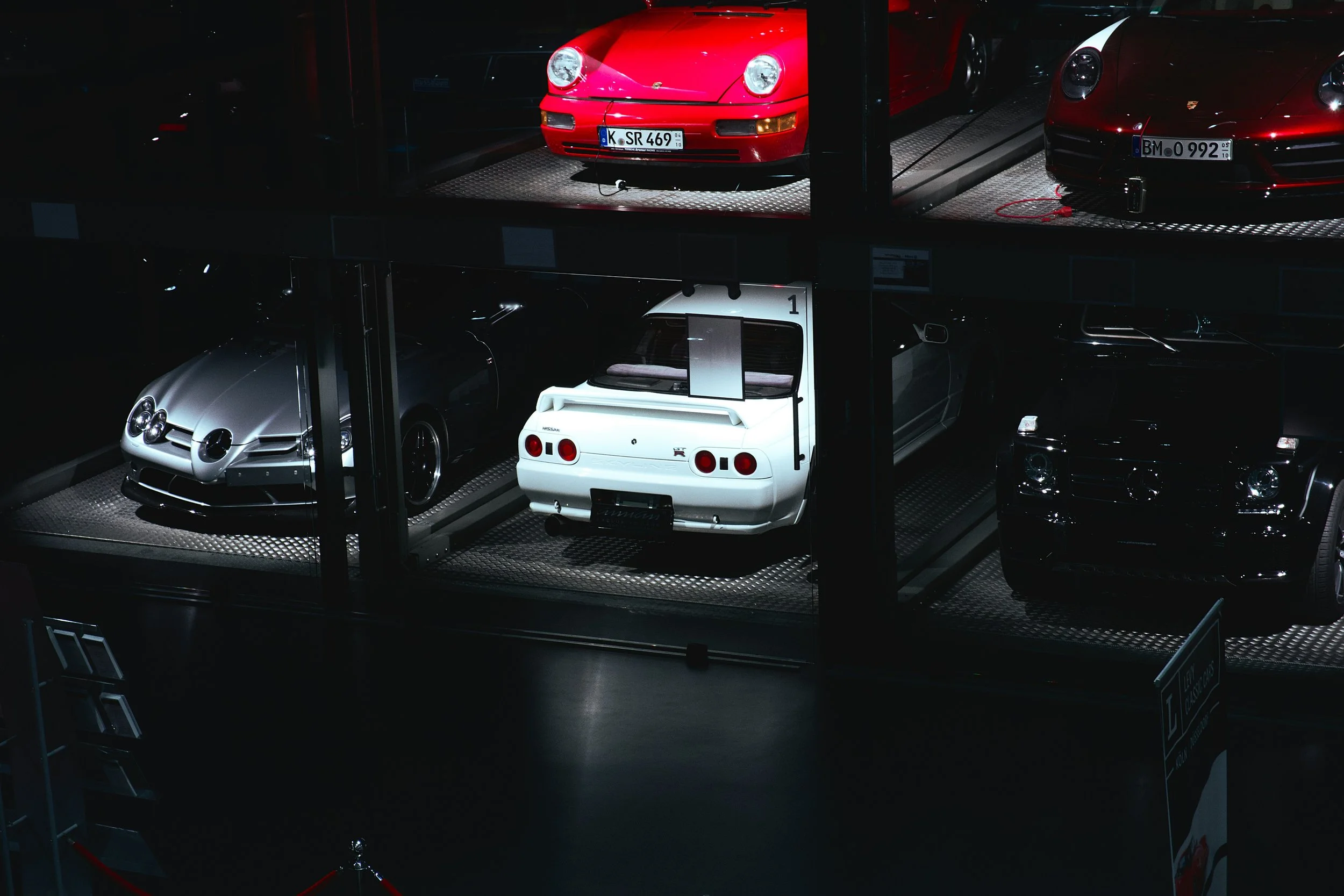 A display of classic and luxury cars in a glass-enclosed showroom, including a red Porsche 911, a silver Mercedes-Benz, a white Nissan Skyline GT-R, and a black Mercedes-Benz G-Class.