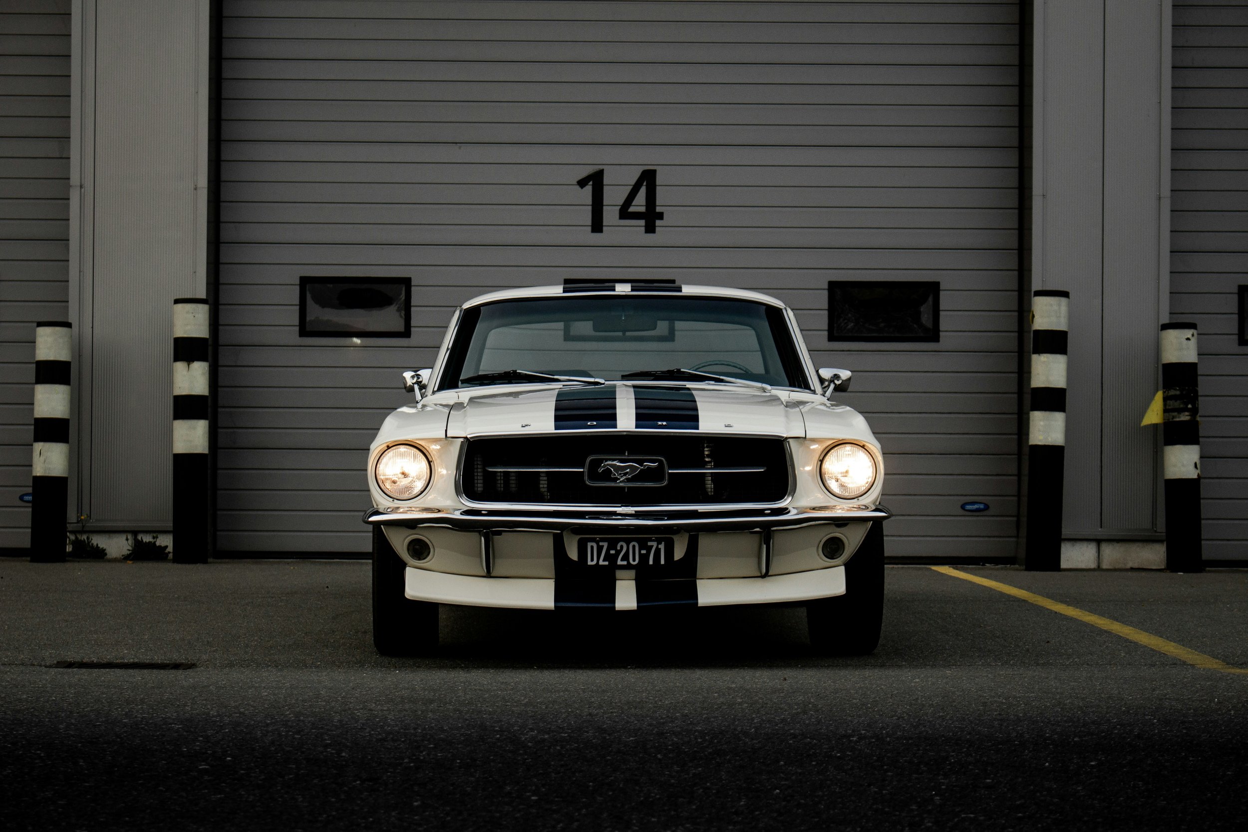A white vintage Ford Mustang with black racing stripes parked in front of a gray garage door numbered 14.