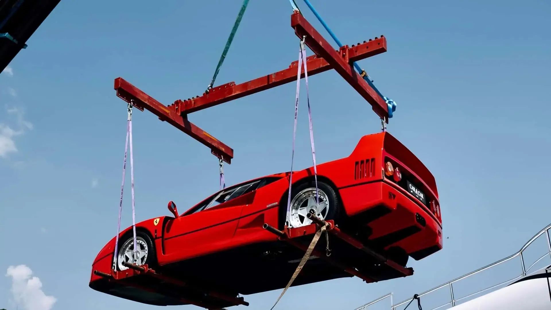 A red Ferrari sports car being lifted by a crane against a blue sky.