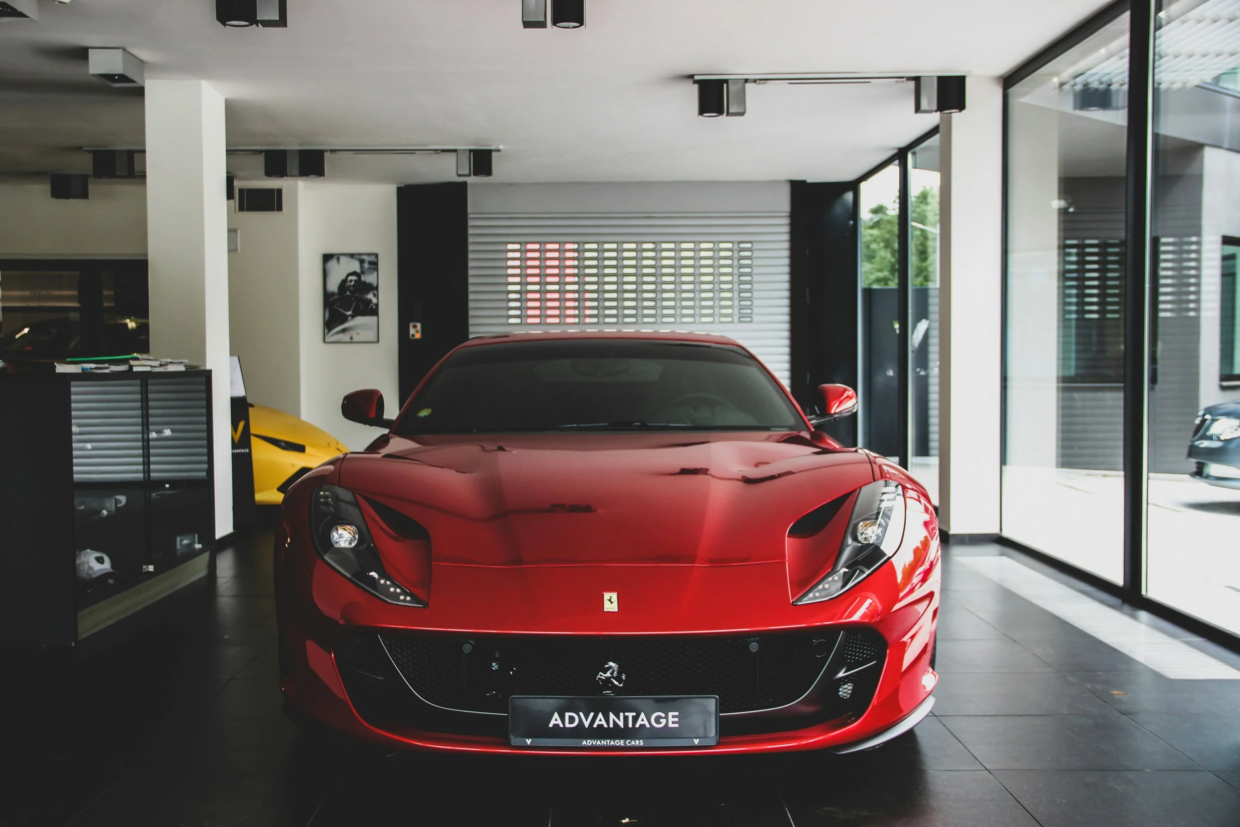Red Ferrari sports car inside a showroom, with black-tiled flooring, large glass windows on the right, and a yellow car partially visible on the left.