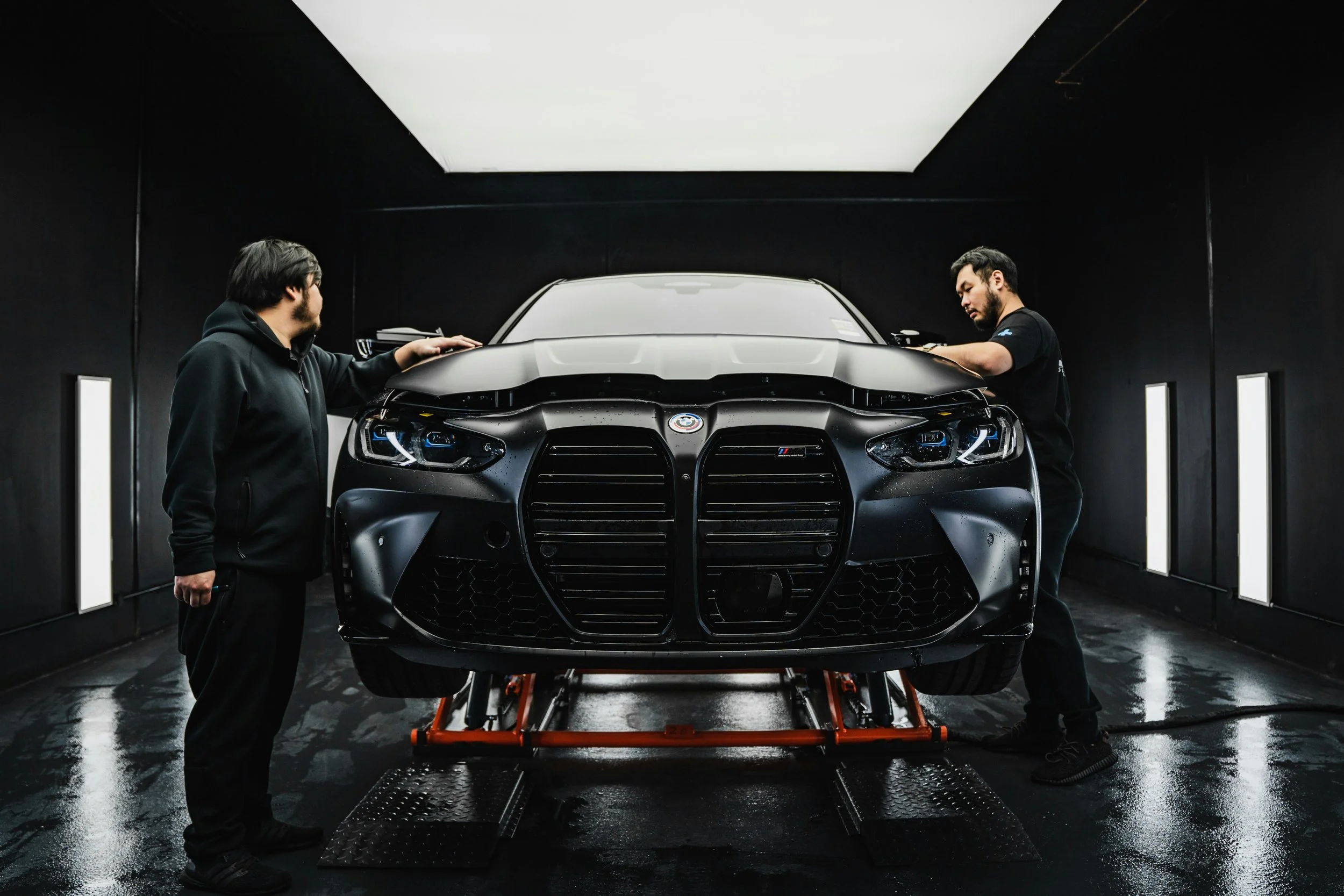 Two mechanics working on a black BMW M car in a dark garage.