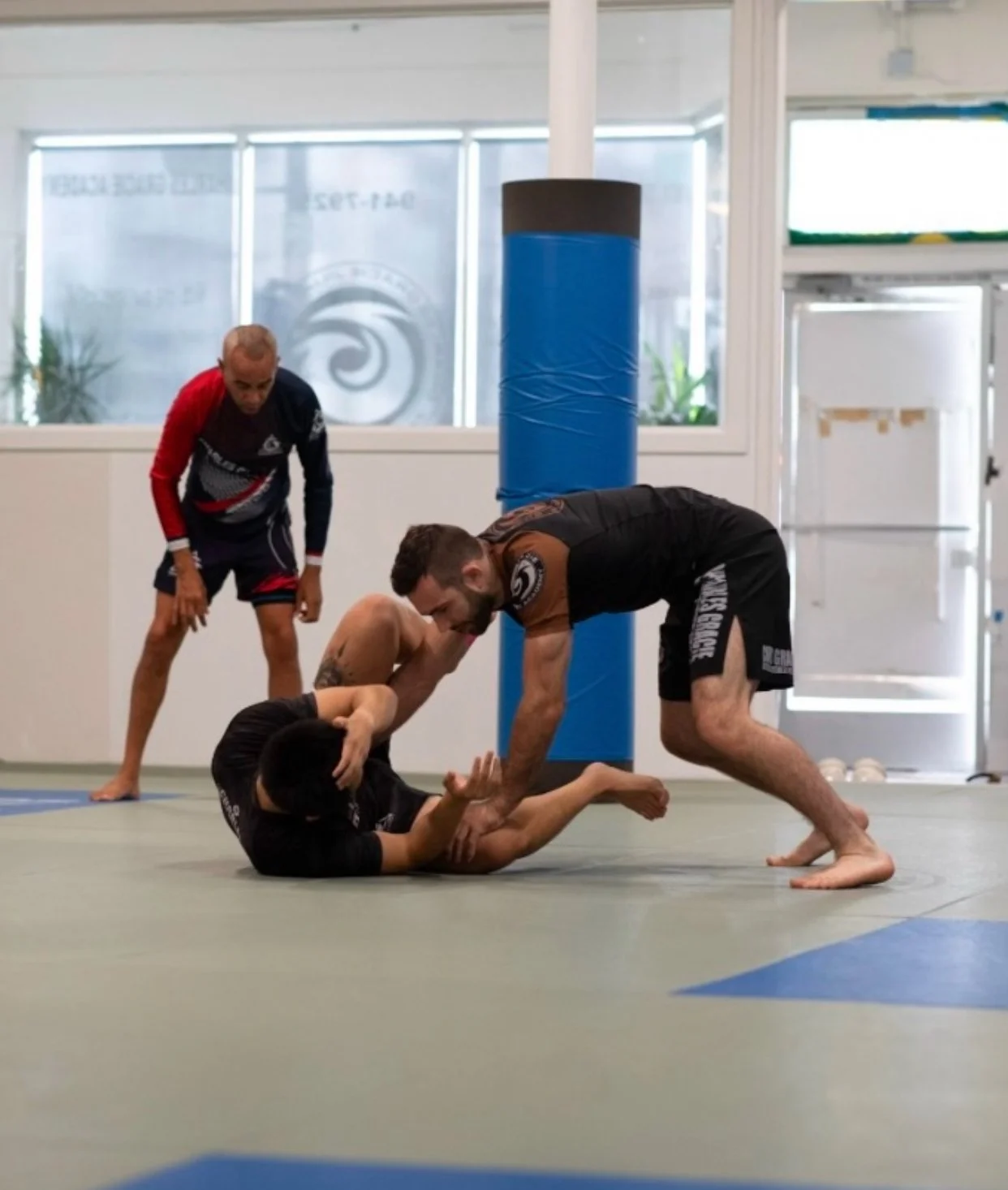 Group of people in a martial arts or boxing training room, standing on mats, some with hands on hips, facing a man in a black shirt who appears to be instructing.