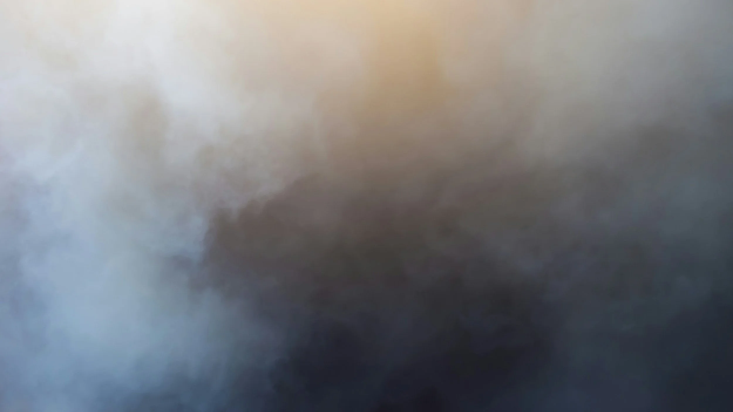 A modern house with wooden and stone exterior walls situated on a hill with green and brown vegetation, with snow-capped mountains in the background and a cloudy sky overhead.
