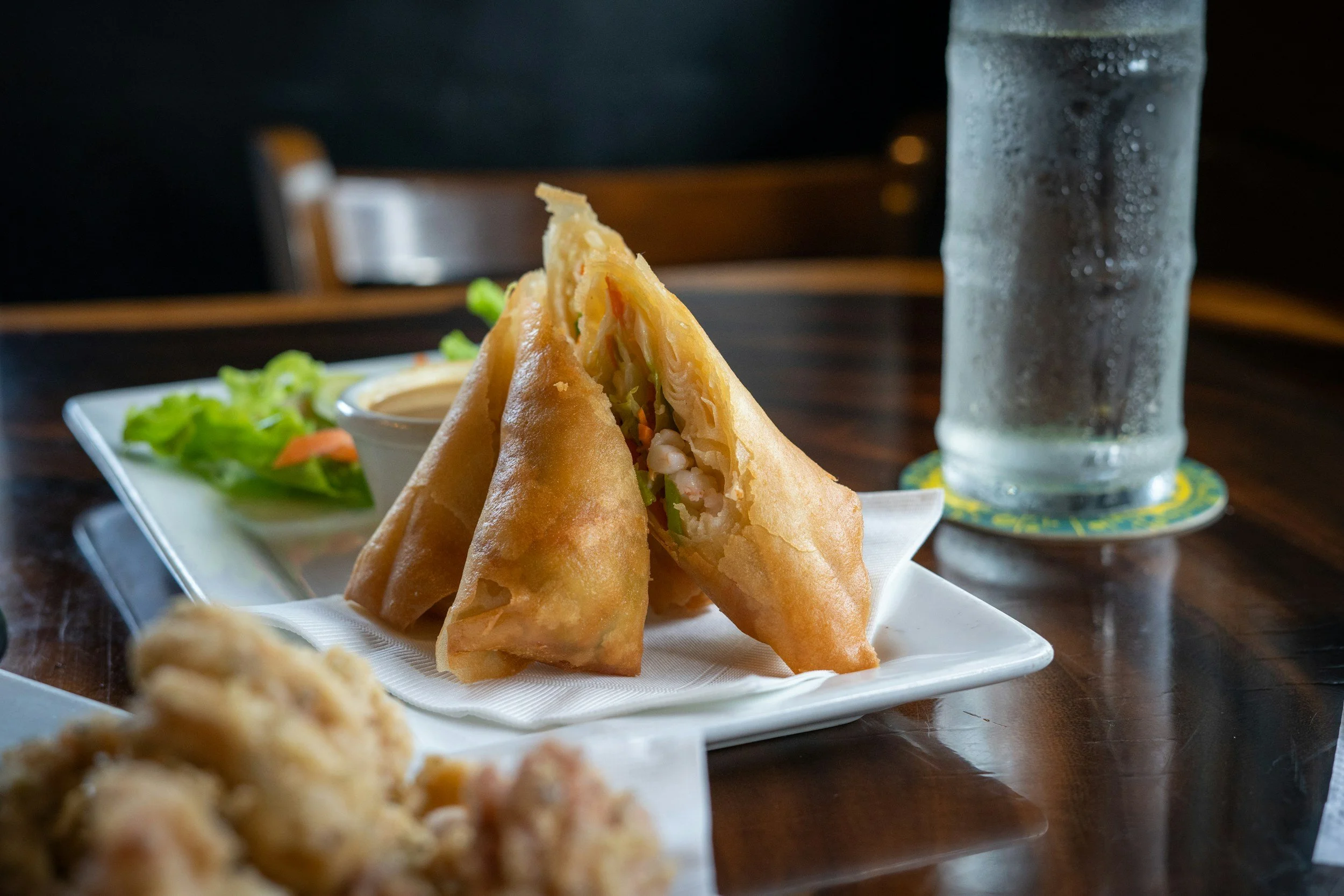 Triangular fried spring rolls on a white plate with lettuce, a small dish of dipping sauce, and a glass of water on a wooden table.