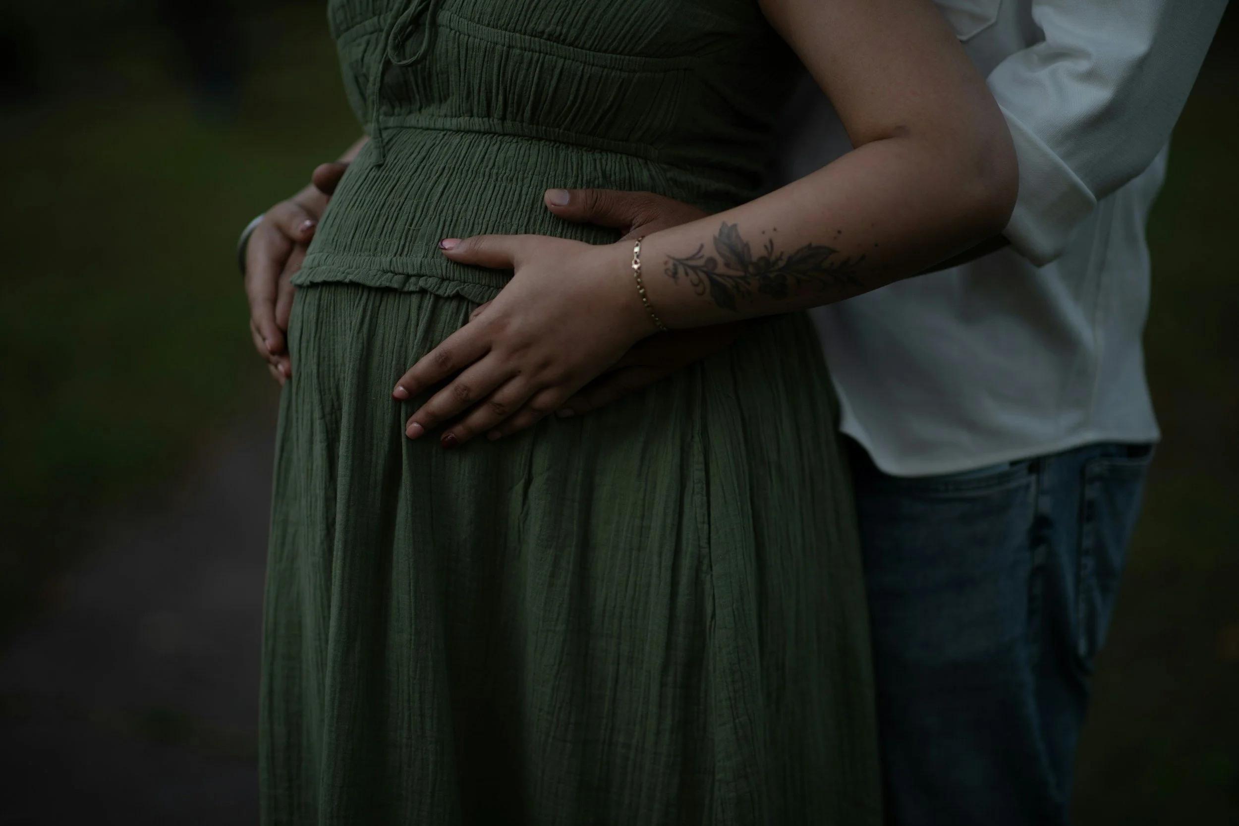 A person wearing a green dress and a tattoo on their arm is being embraced from behind by another person, with hands gently placed on the person's pregnant belly.