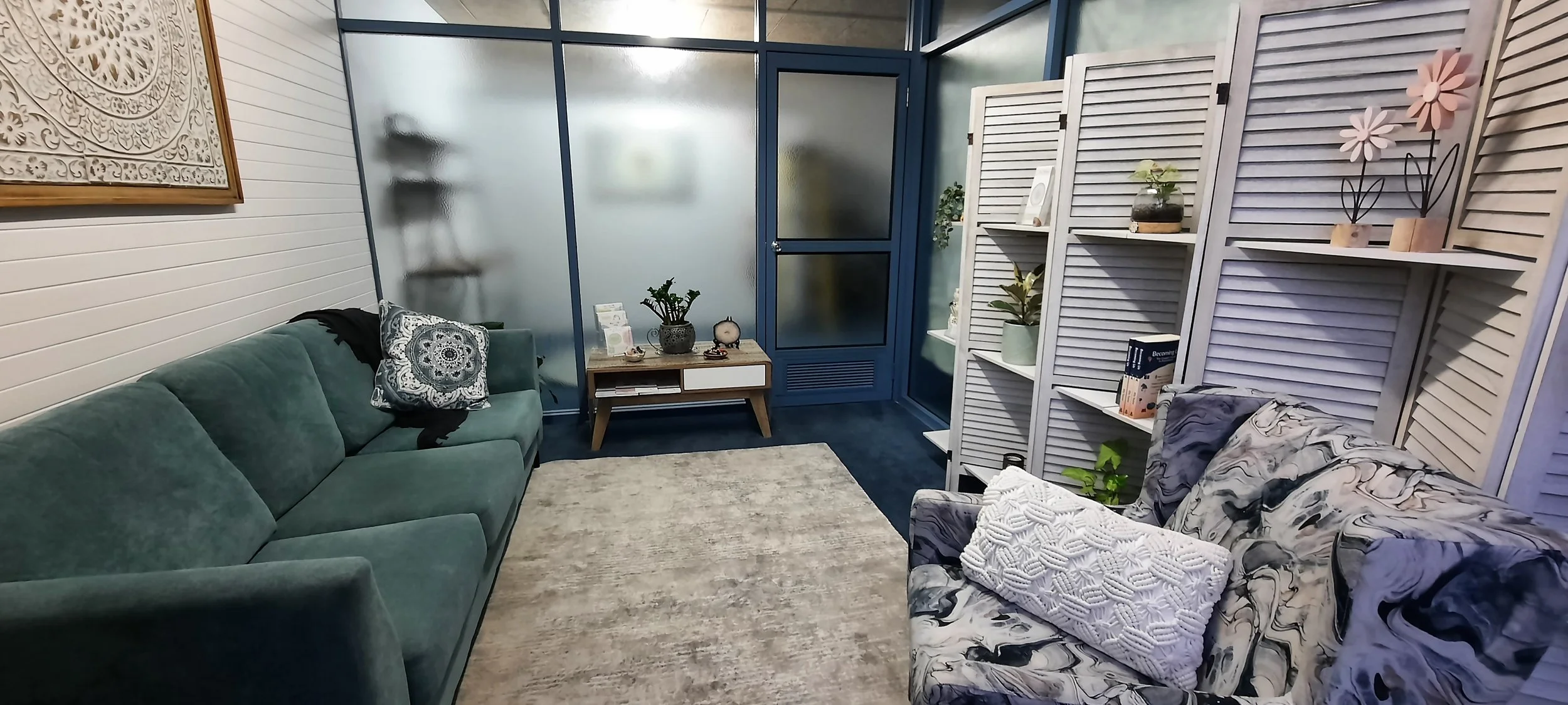Living room with a green sofa, patterned pillows, a beige rug, a wooden side table, decorative plants, white louvered room dividers, and a window with frosted glass.