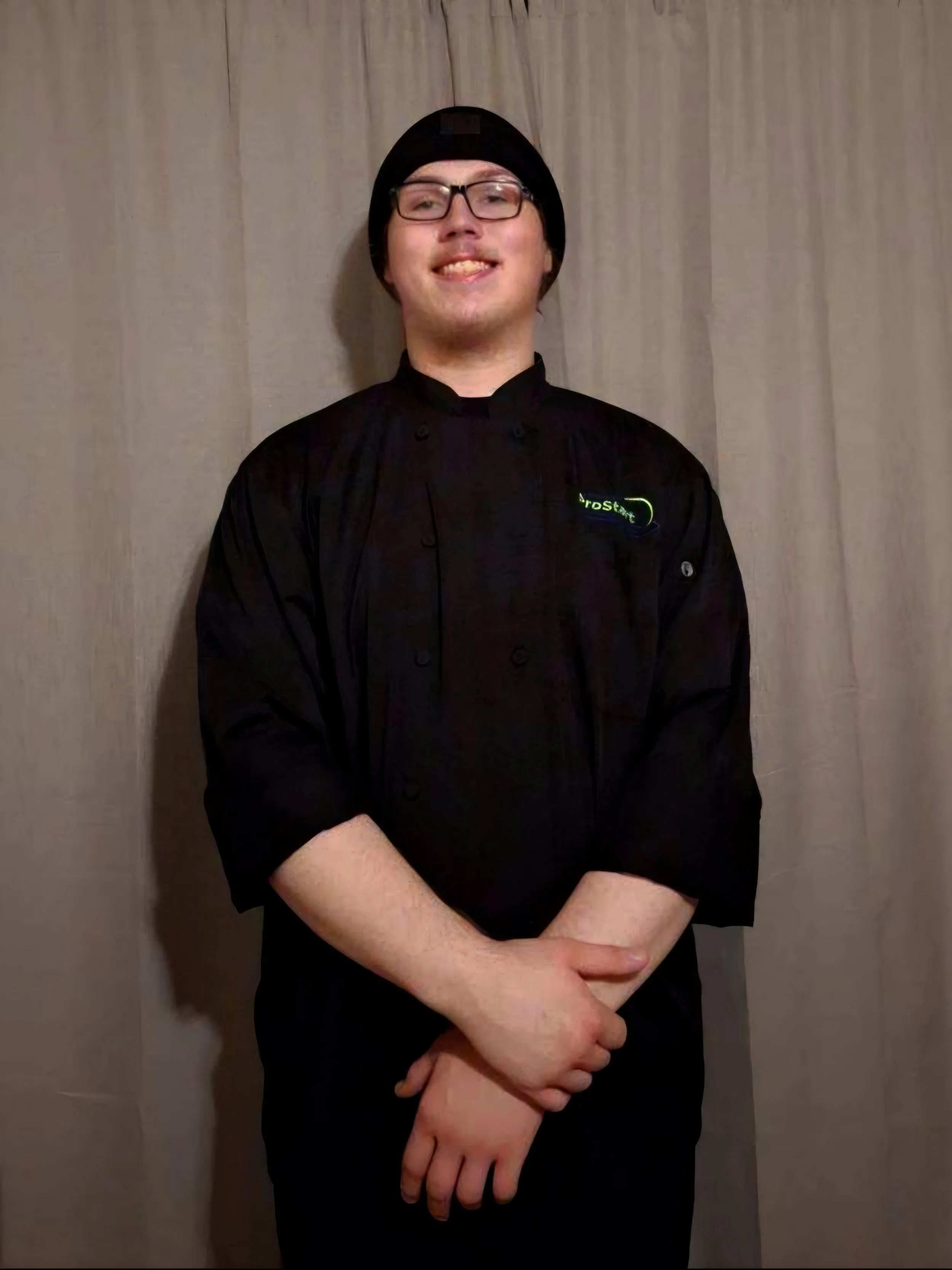 A smiling person wearing glasses, a black chef's coat, and a black beanie, standing against a beige curtain background.