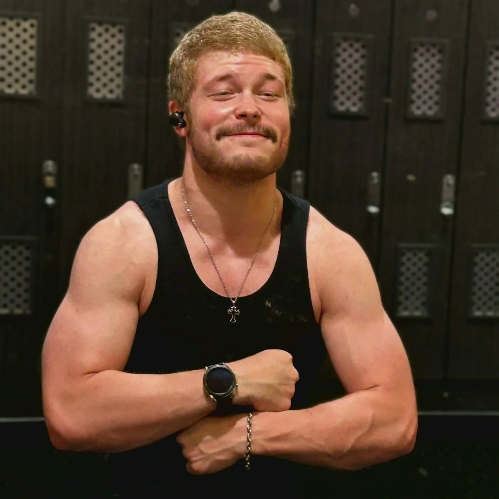 A muscular man with red hair and a beard, smiling confidently, wearing a black sleeveless shirt, a necklace with a cross pendant, a watch, and a bracelet, standing in front of dark lockers.