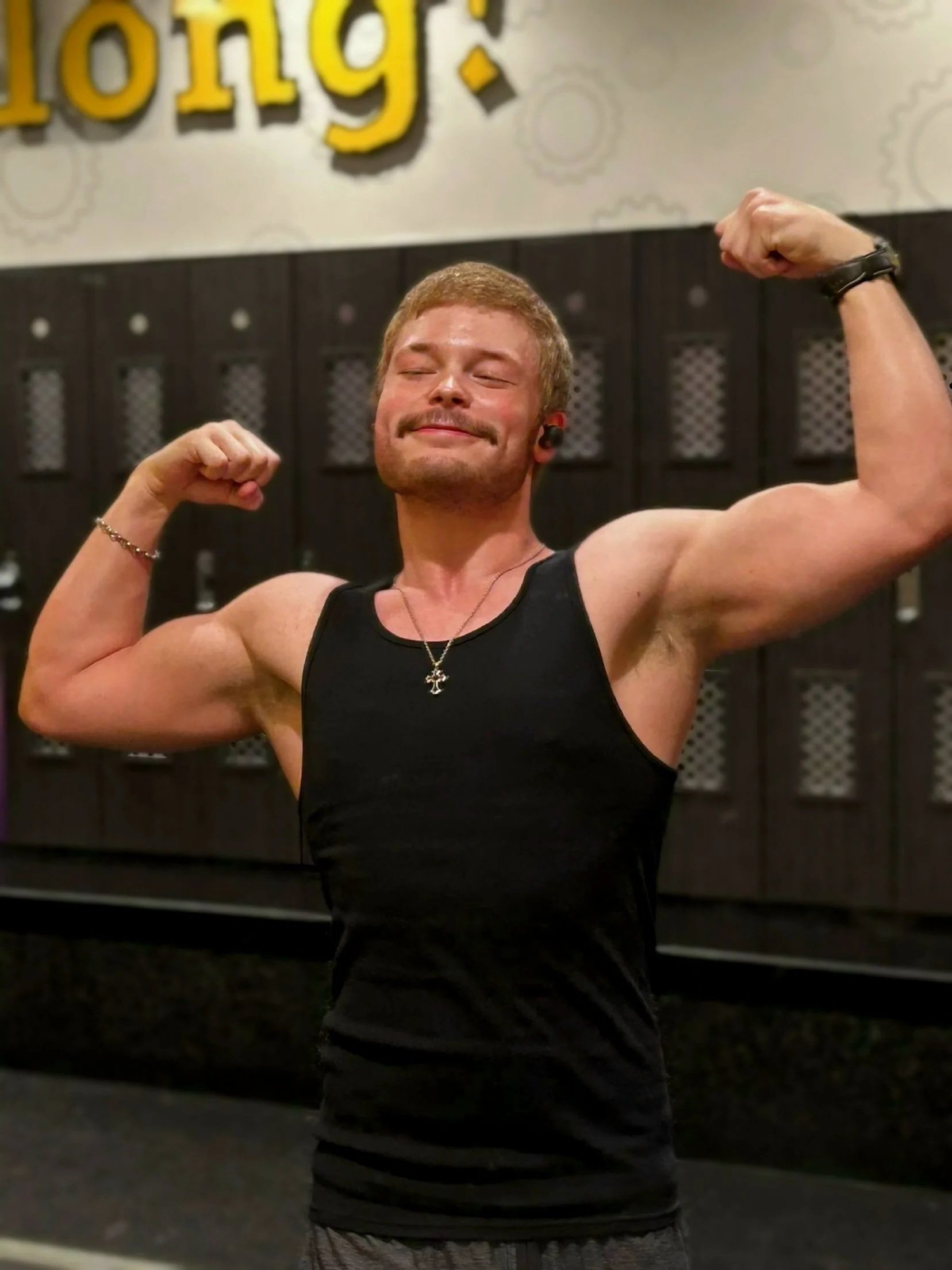 Young man flexing his biceps in a gym, smiling with eyes closed, wearing a black sleeveless shirt, necklace, and smartwatch, with gym lockers in the background.