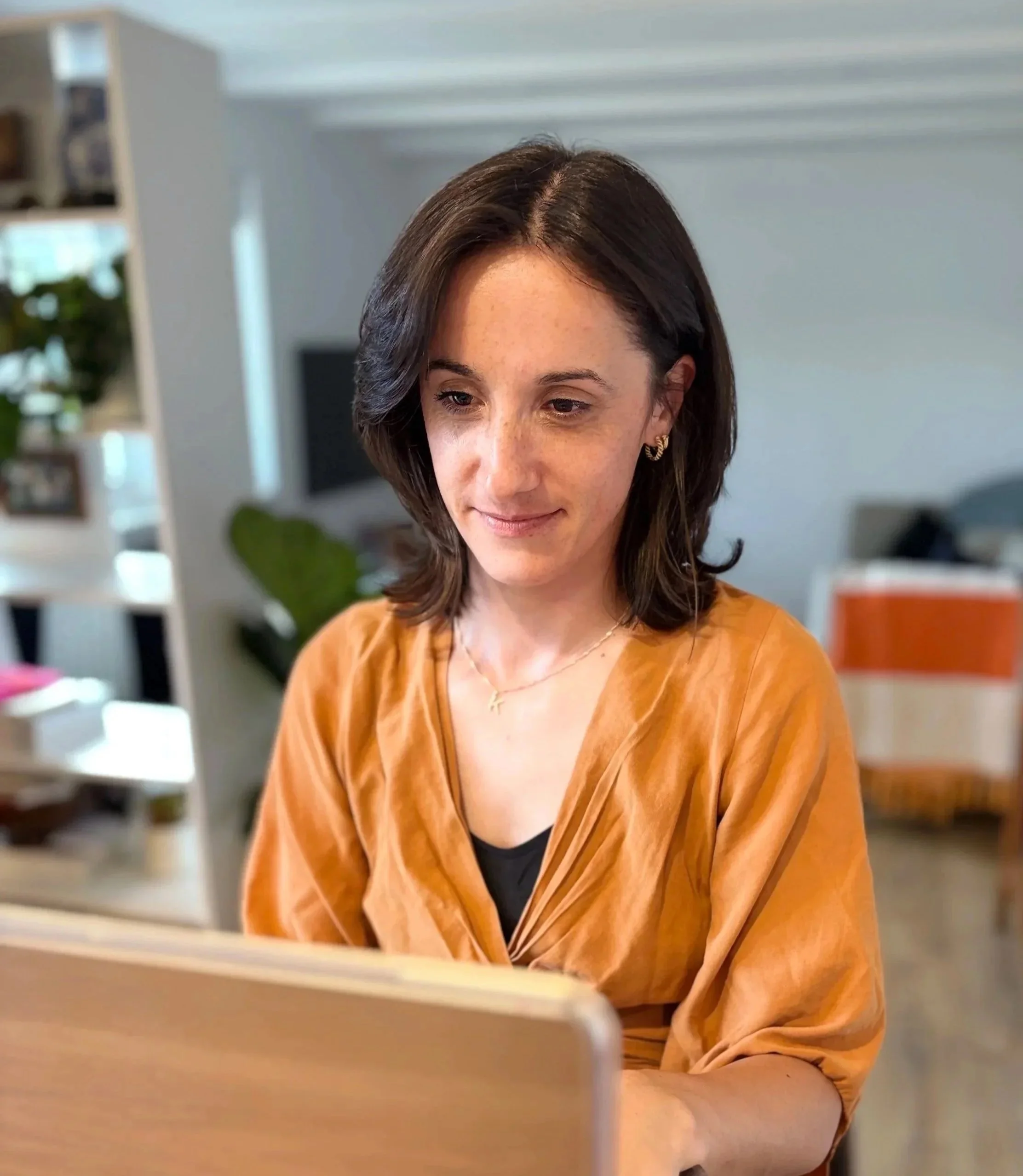 A woman with shoulder-length dark hair, wearing a rust-colored blouse and gold jewelry, sitting and looking at a laptop.