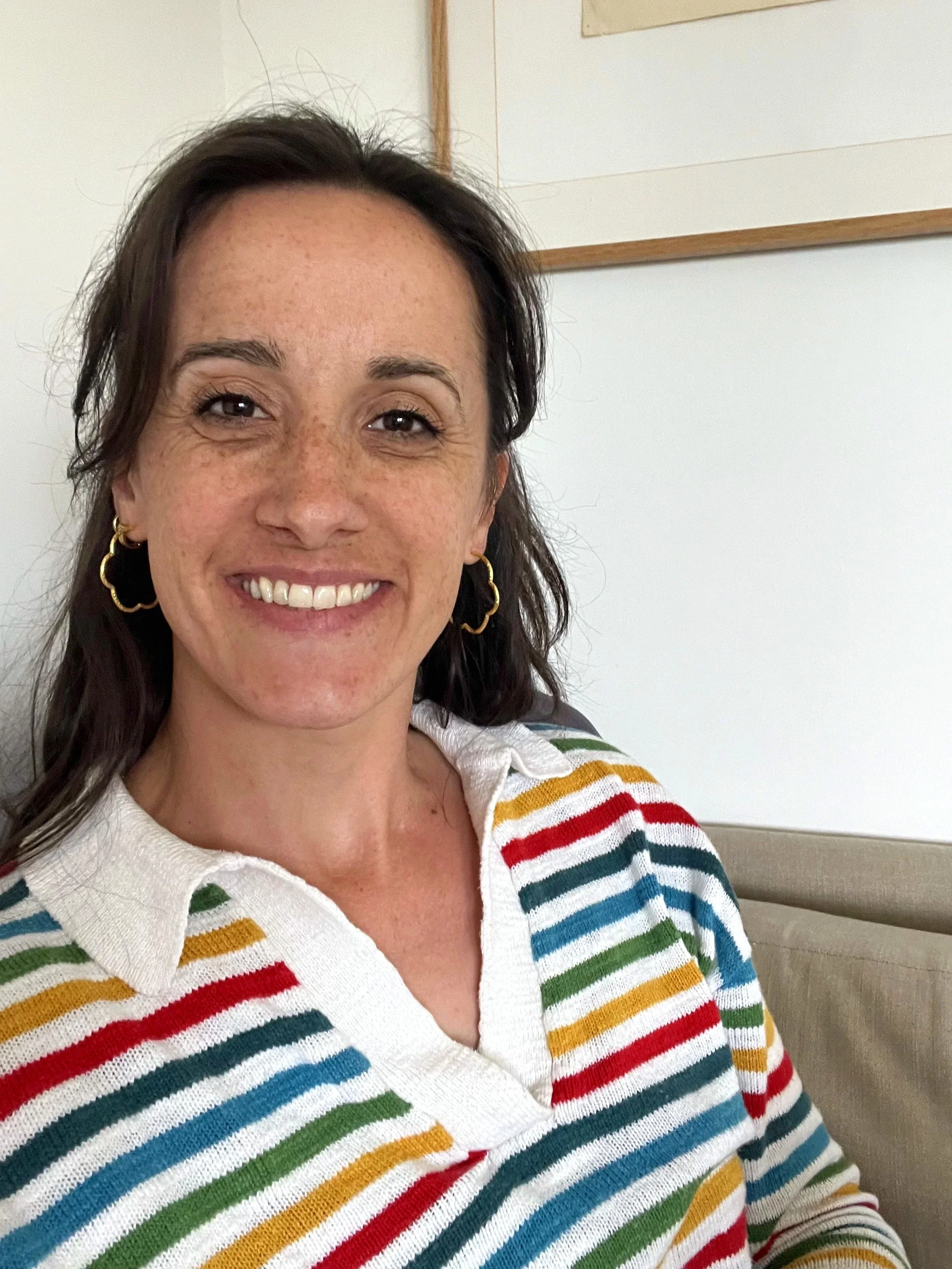 A woman smiling, wearing gold hoop earrings and a colorful striped sweater, sitting in a room with white walls and a wooden frame on the wall.