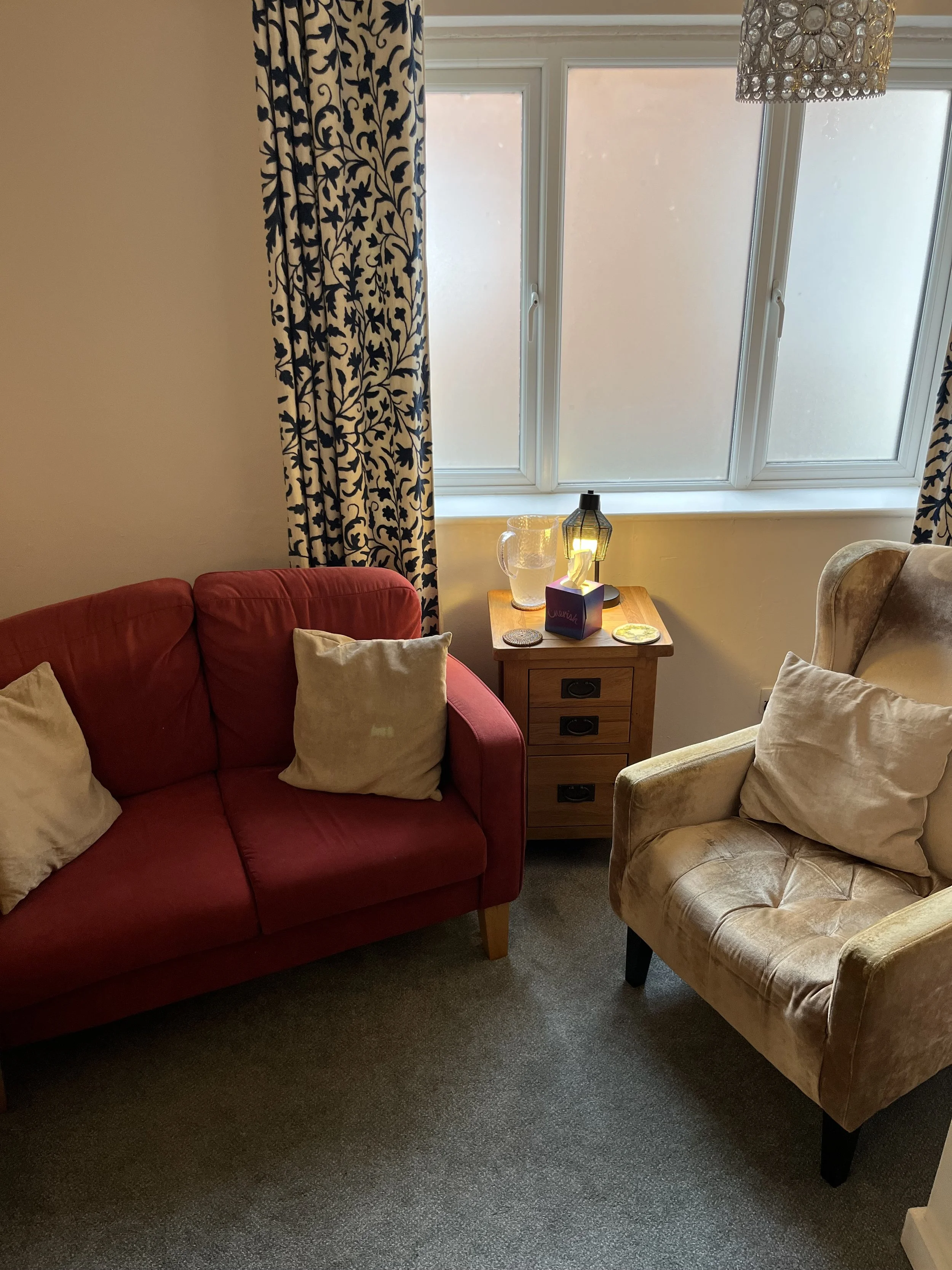 A cozy living room corner with a red sofa and beige armchair, a wooden side table with a lamp, pitcher, and decorative objects, large window with blue and white floral curtains, and a modern chandelier above.